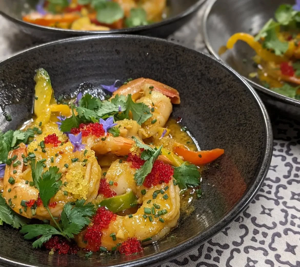 Close-up of a black bowl of shrimp and vegetable stir-fry garnished with cilantro, edible flowers, and red roe, with other similar dishes partially visible in the background.