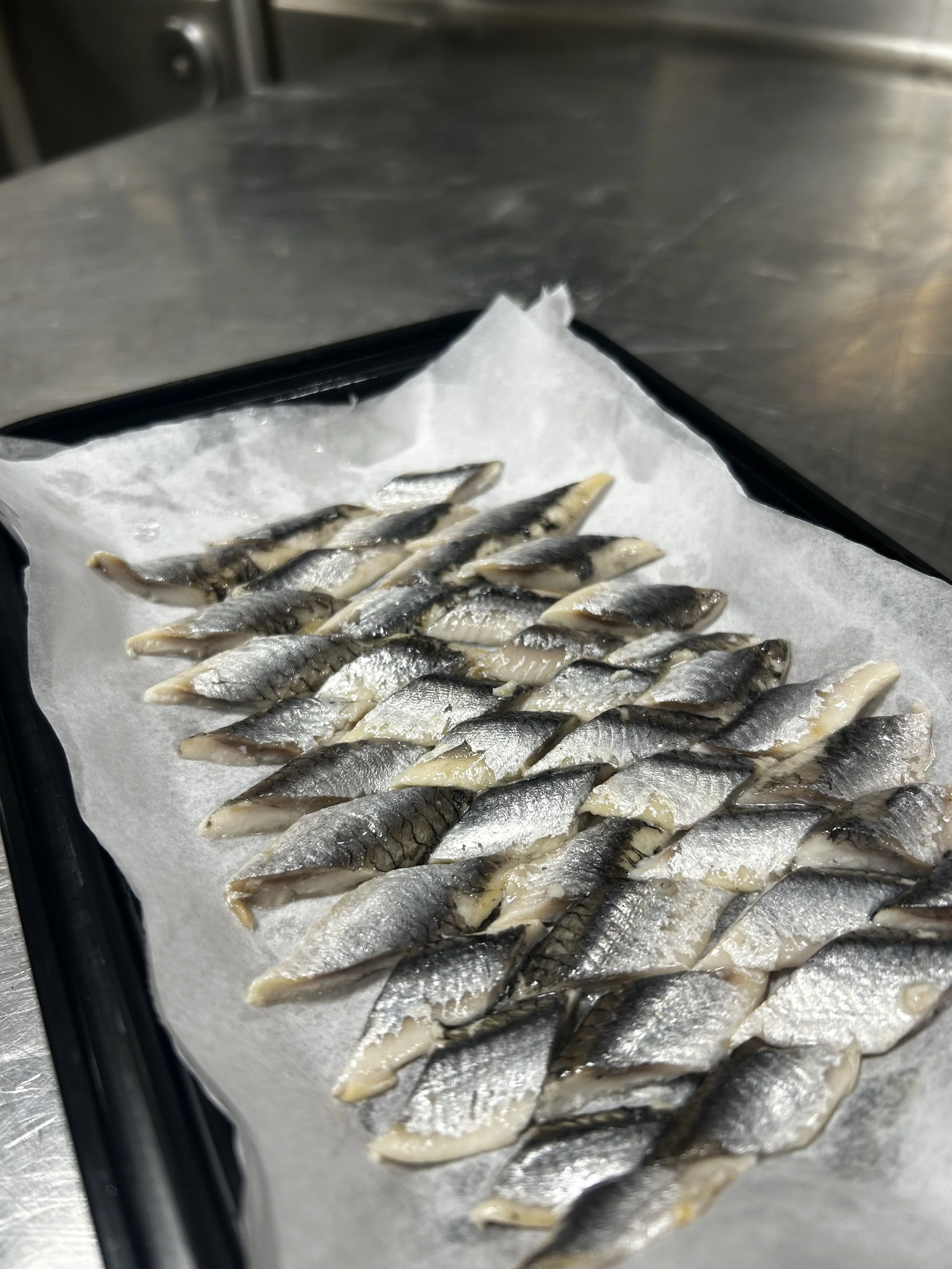 Small fish fillets lined up on parchment paper on a baking tray in a kitchen or food preparation area.