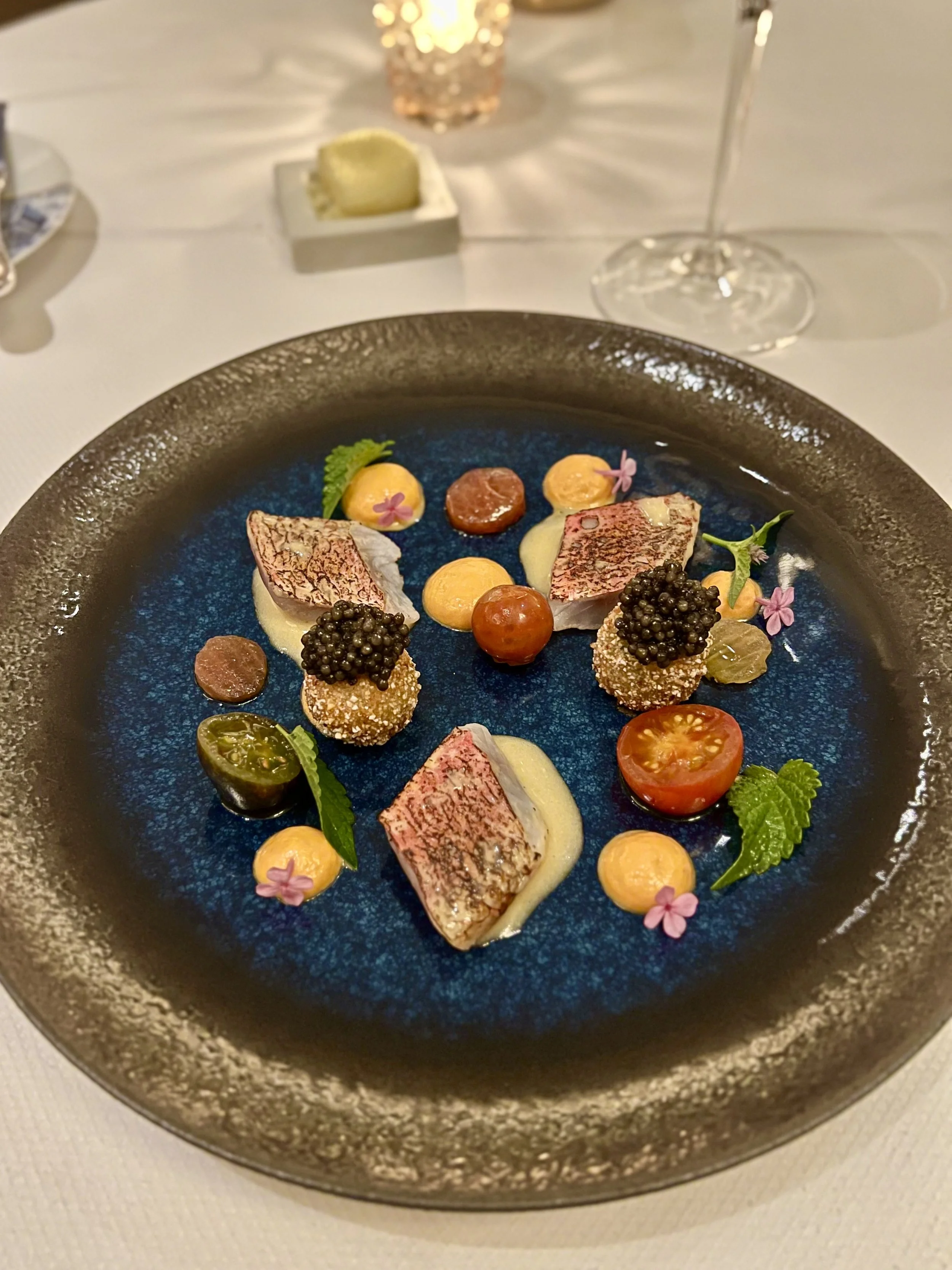A gourmet dish featuring slices of cooked fish, cherry tomatoes, caviar, small dollops of sauce, green leaves, and edible flowers on a dark, textured plate.