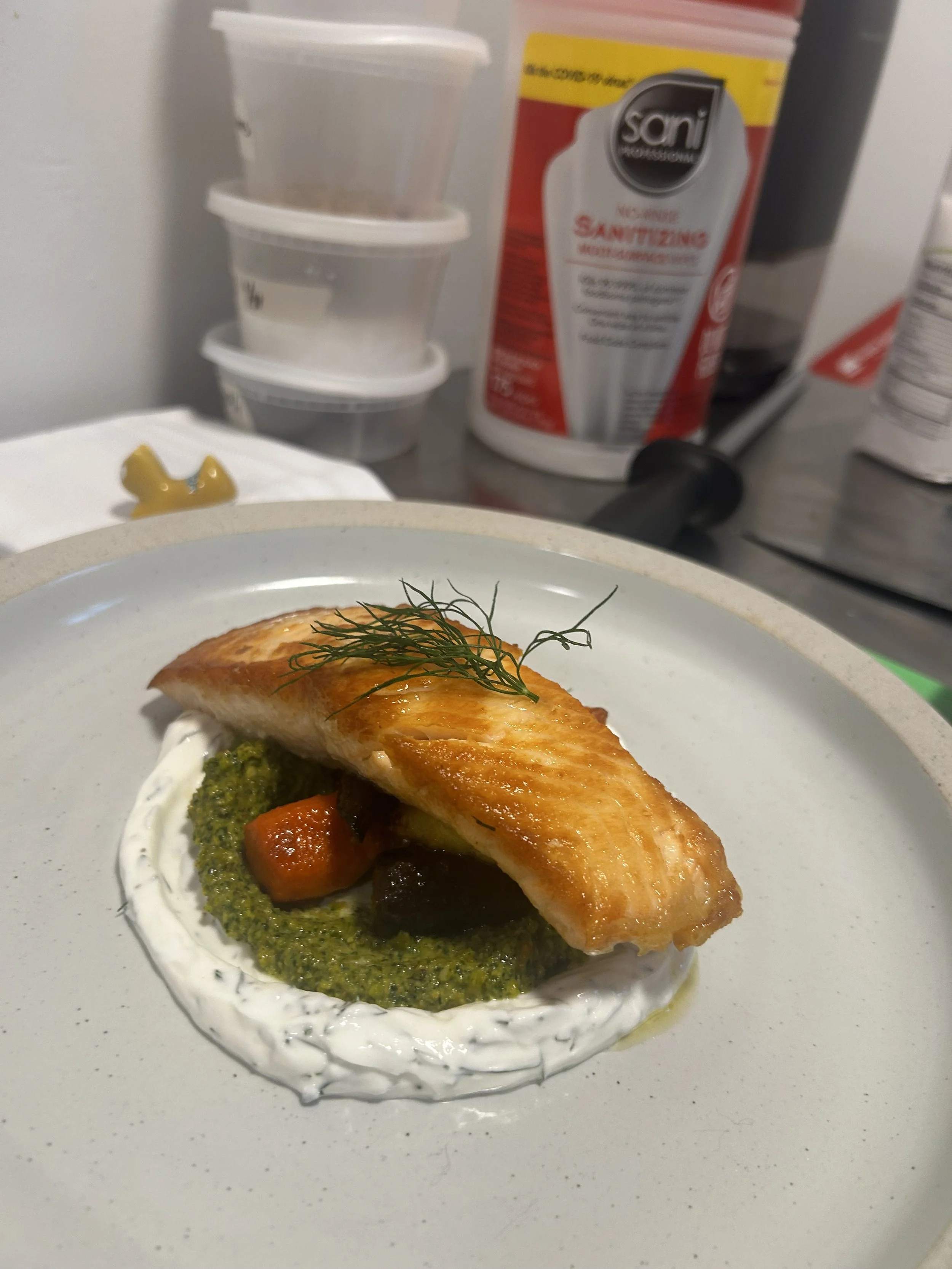 A plated dish featuring a piece of cooked fish resting on a bed of green sauce, accompanied by small vegetables, topped with dill garnish.