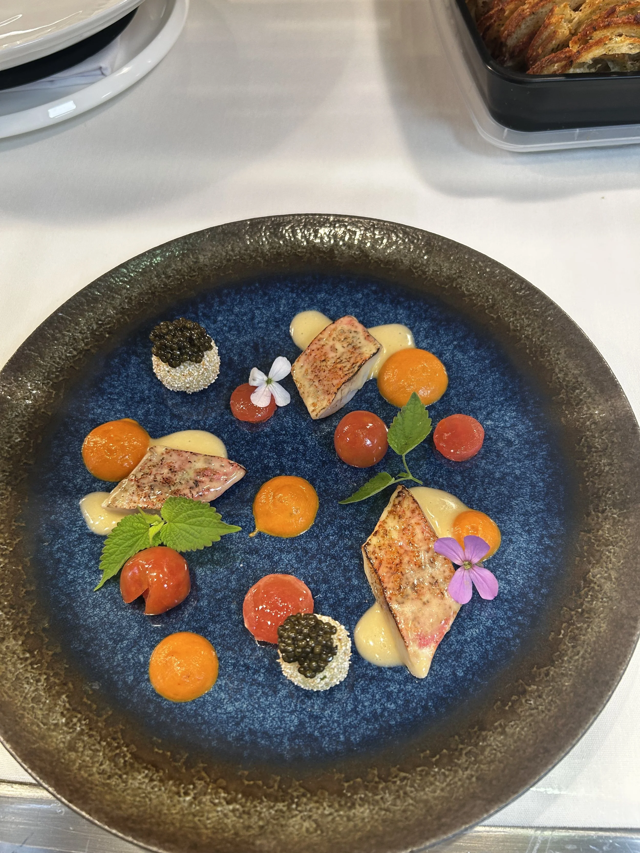 An elegantly plated dish featuring seared fish, small dollops of sauce, colorful garnishes, edible flowers, and caviar on a dark textured plate.