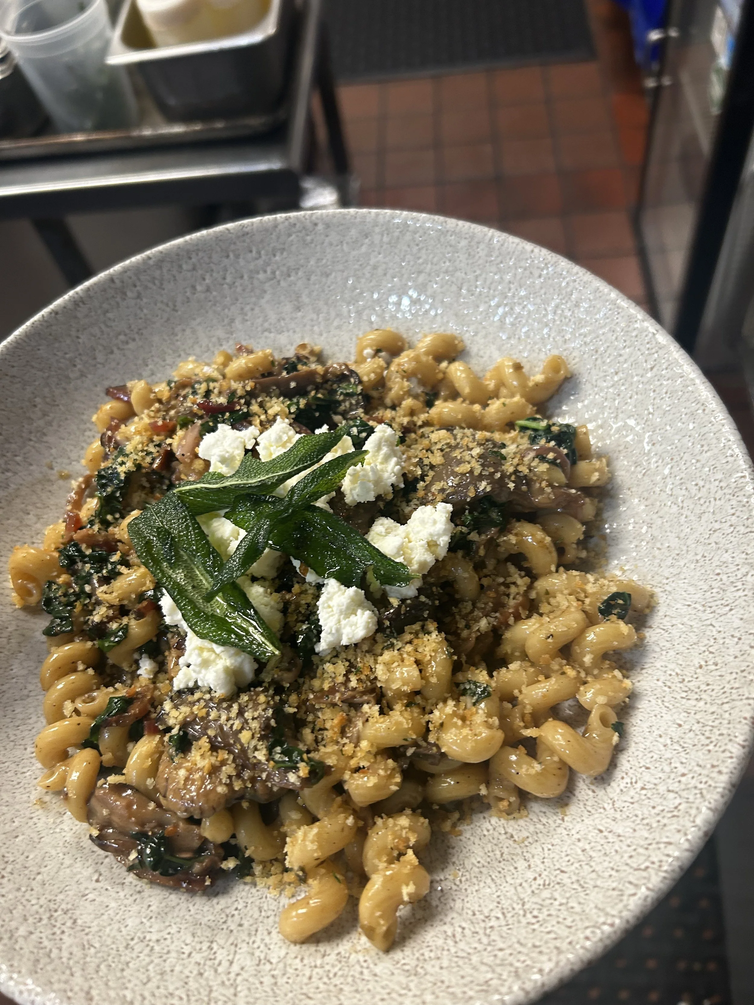 Close-up of a bowl of pasta with greens, grated cheese, and breadcrumb topping, in a restaurant setting.