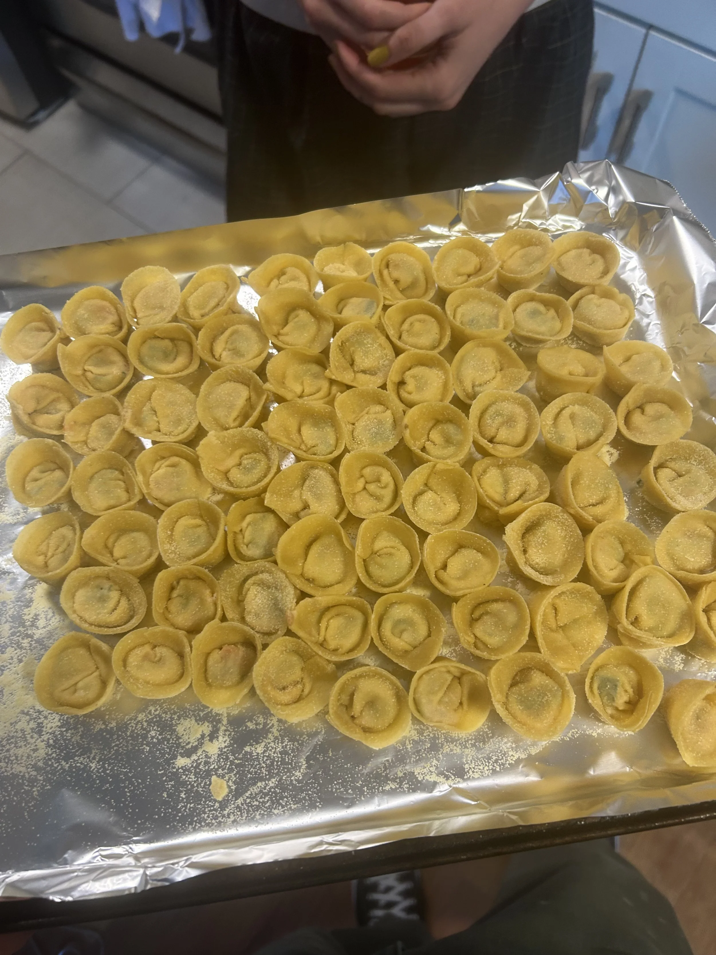 Unbaked tortellini pasta on a baking sheet covered with aluminum foil, ready for cooking, with a person in the background.