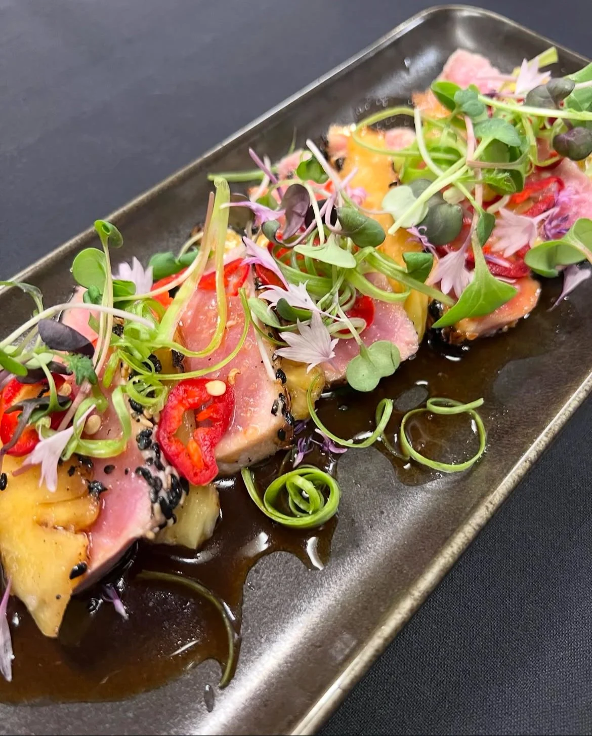Sliced raw fish topped with microgreens, red pepper slices, and black sesame seeds on a black rectangular plate with soy sauce.