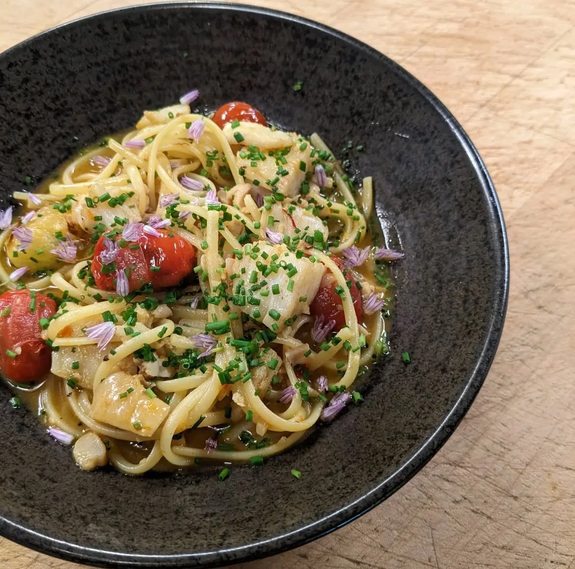 Pasta dish with cherry tomatoes, chopped chives, and edible flowers in a black bowl on a wooden surface.