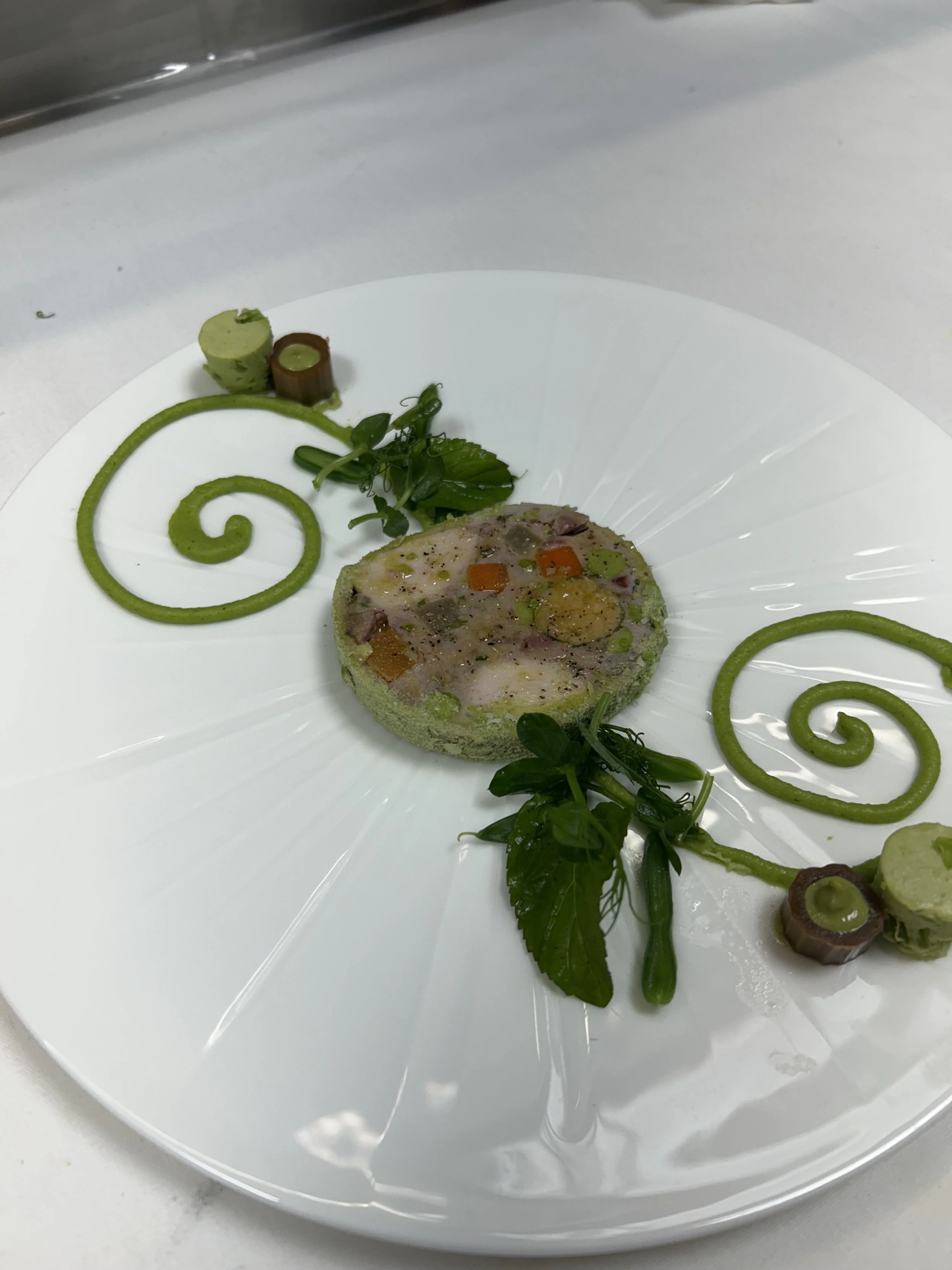 A gourmet dish featuring a confit terrine with vegetables, garnished with green spiral sauces, herbs, and small vegetable rolls on a white plate.