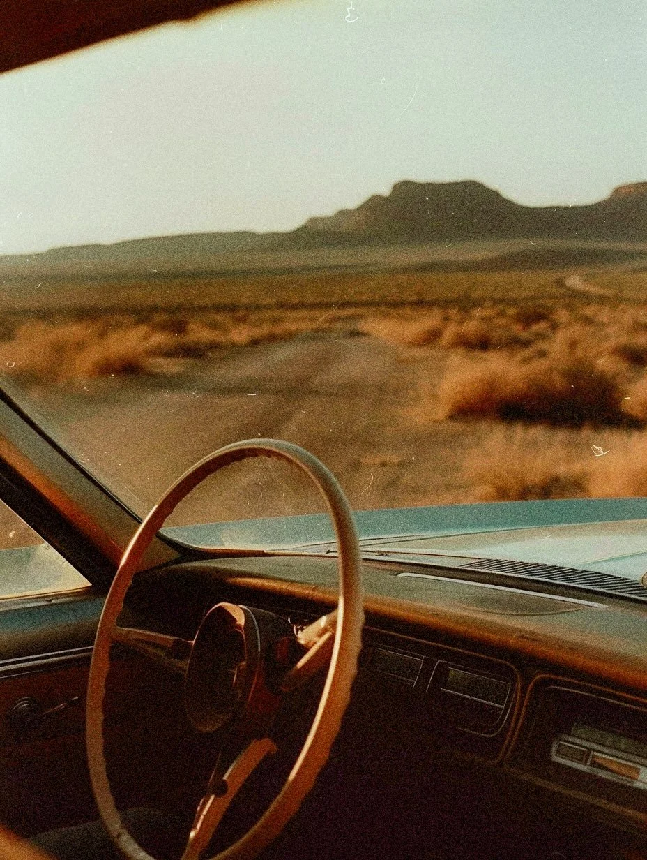 Photo of a car driving on a desert road with mountains in the background, taken from inside the vehicle showing the steering wheel and dashboard.