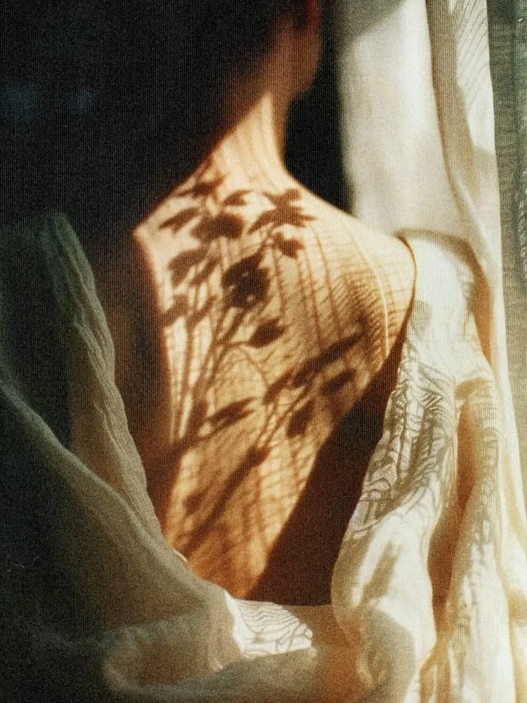 A person sitting by a window with sunlight casting shadow of leaves onto their shoulder and upper arm.