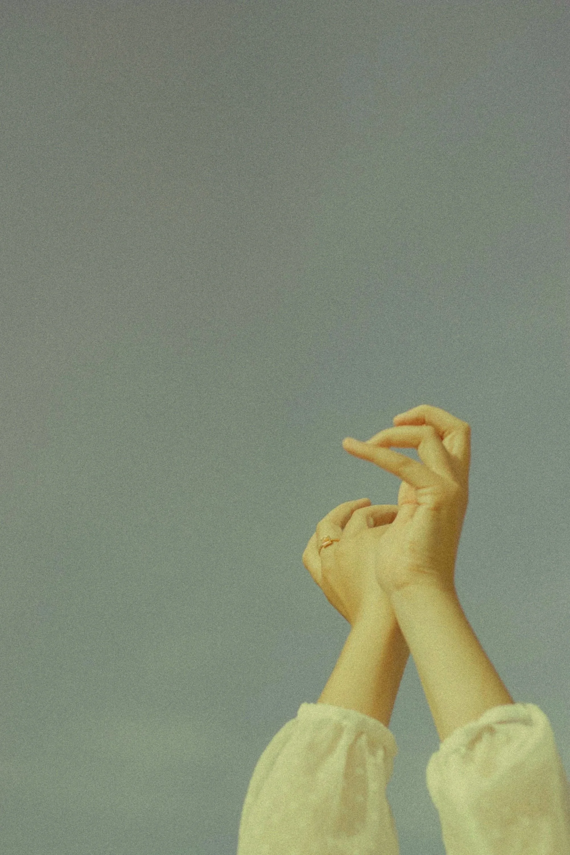 A person with light skin tone making a delicate hand gesture against a plain, muted background. The person wears a lacy, cream-colored garment.