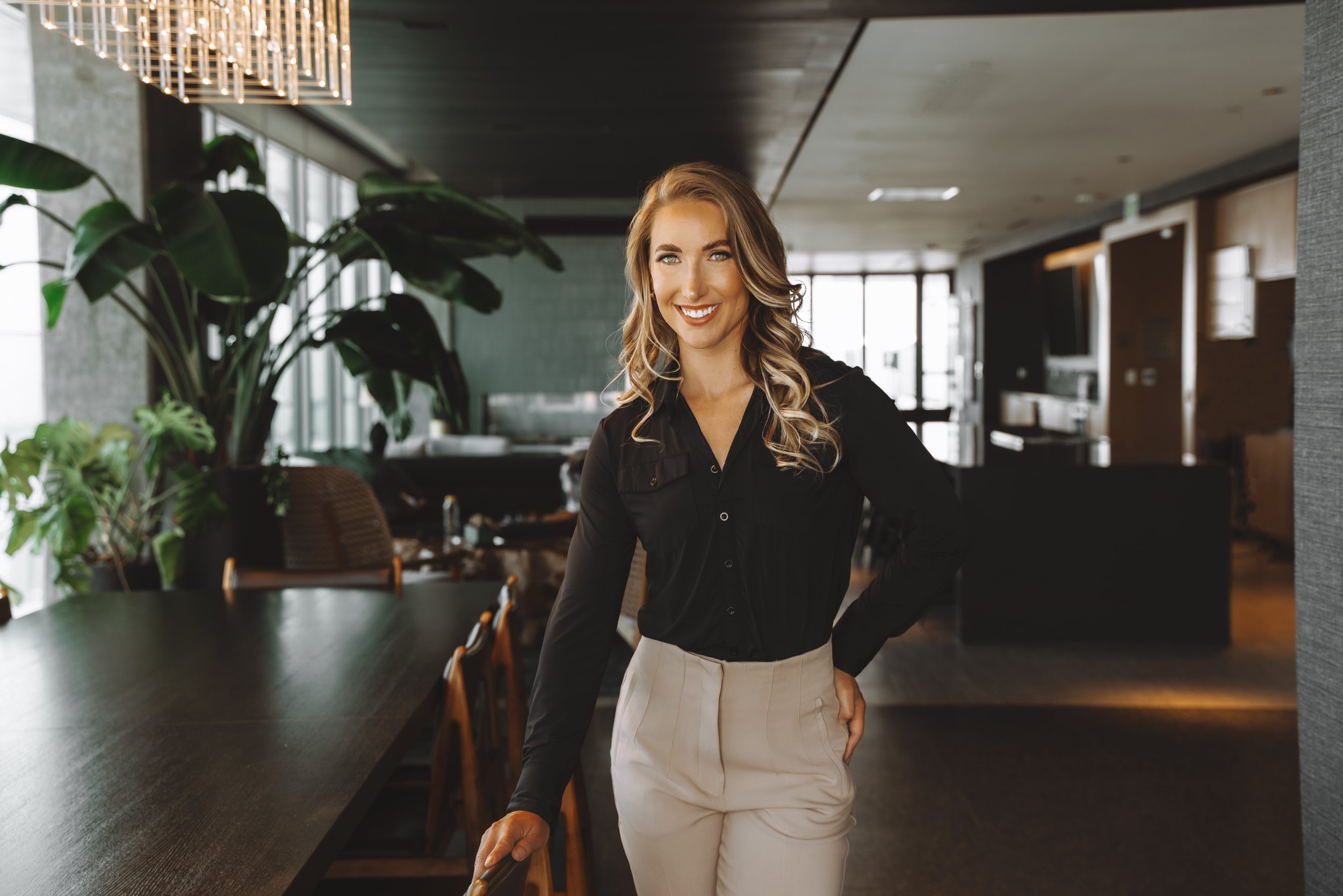 A smiling woman with long wavy hair, wearing a black blouse and beige skirt, standing in a modern restaurant or cafe with large plants and contemporary decor.