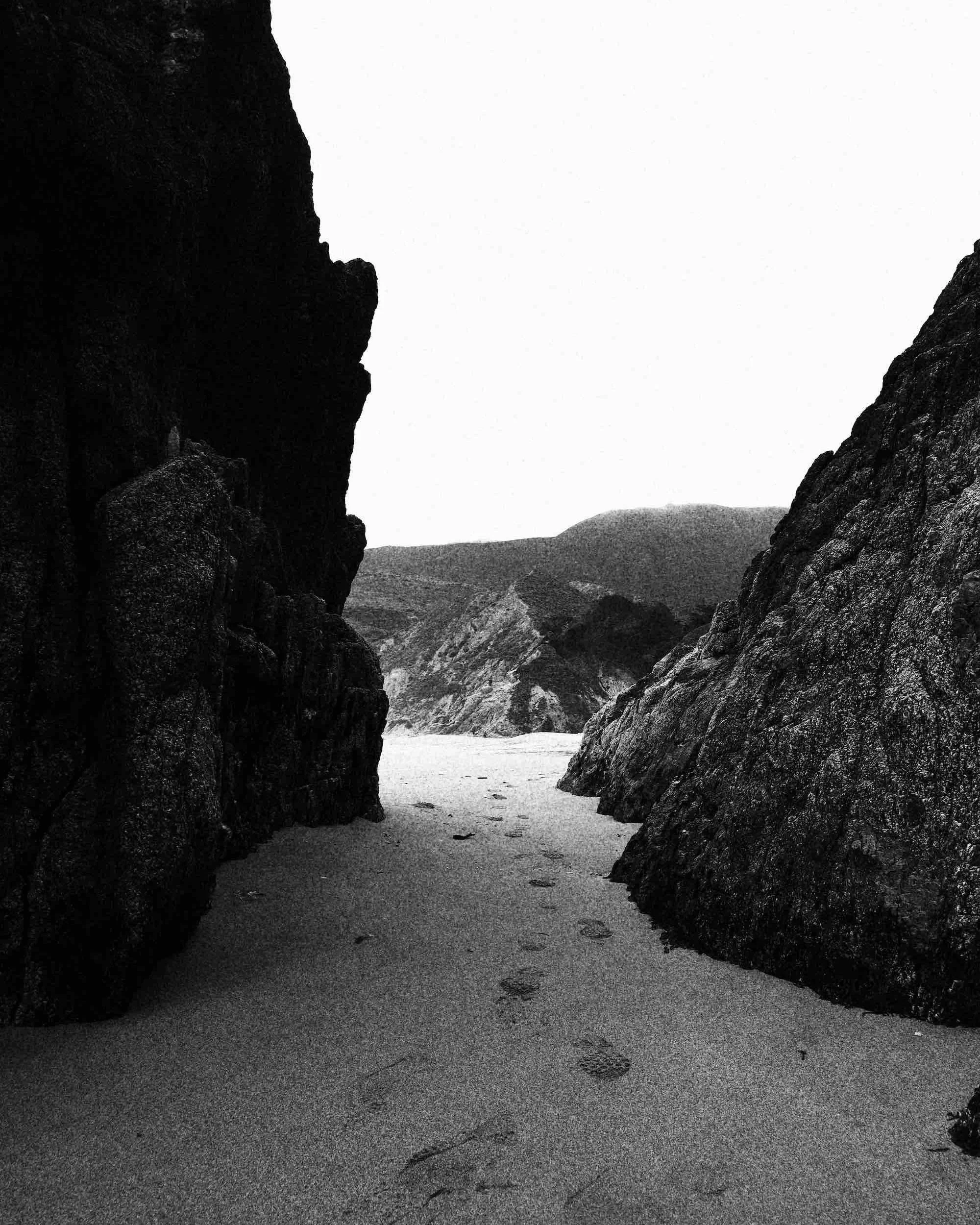 Black and white photo of a sandy beach between dark rock formations with mountains in the background.