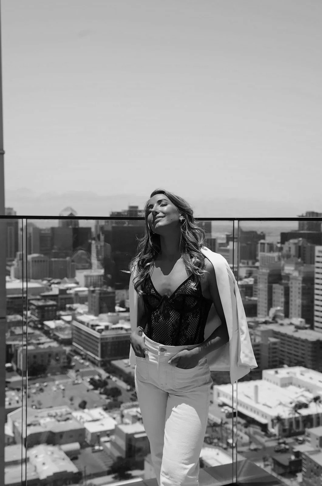 A woman with long wavy hair standing on a balcony with a city skyline in the background, smiling with eyes closed, in black and white.