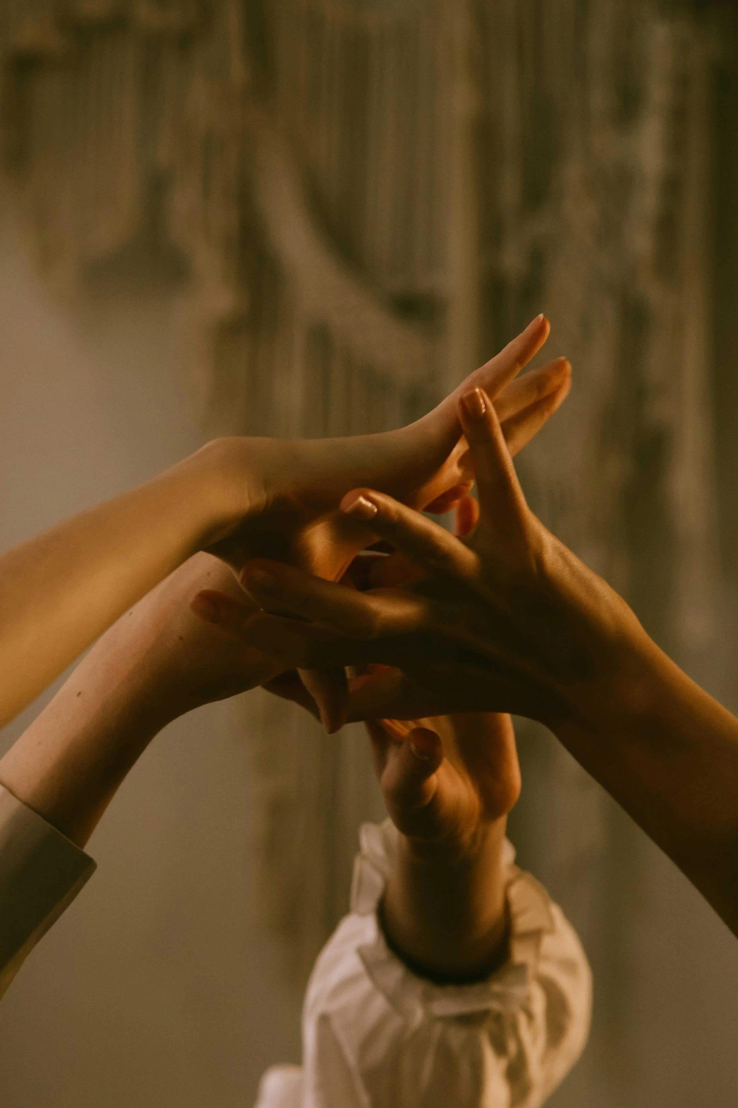 Multiple hands of different skin tones reaching upward with fingers interlaced in a gesture of connection, against a blurred background.