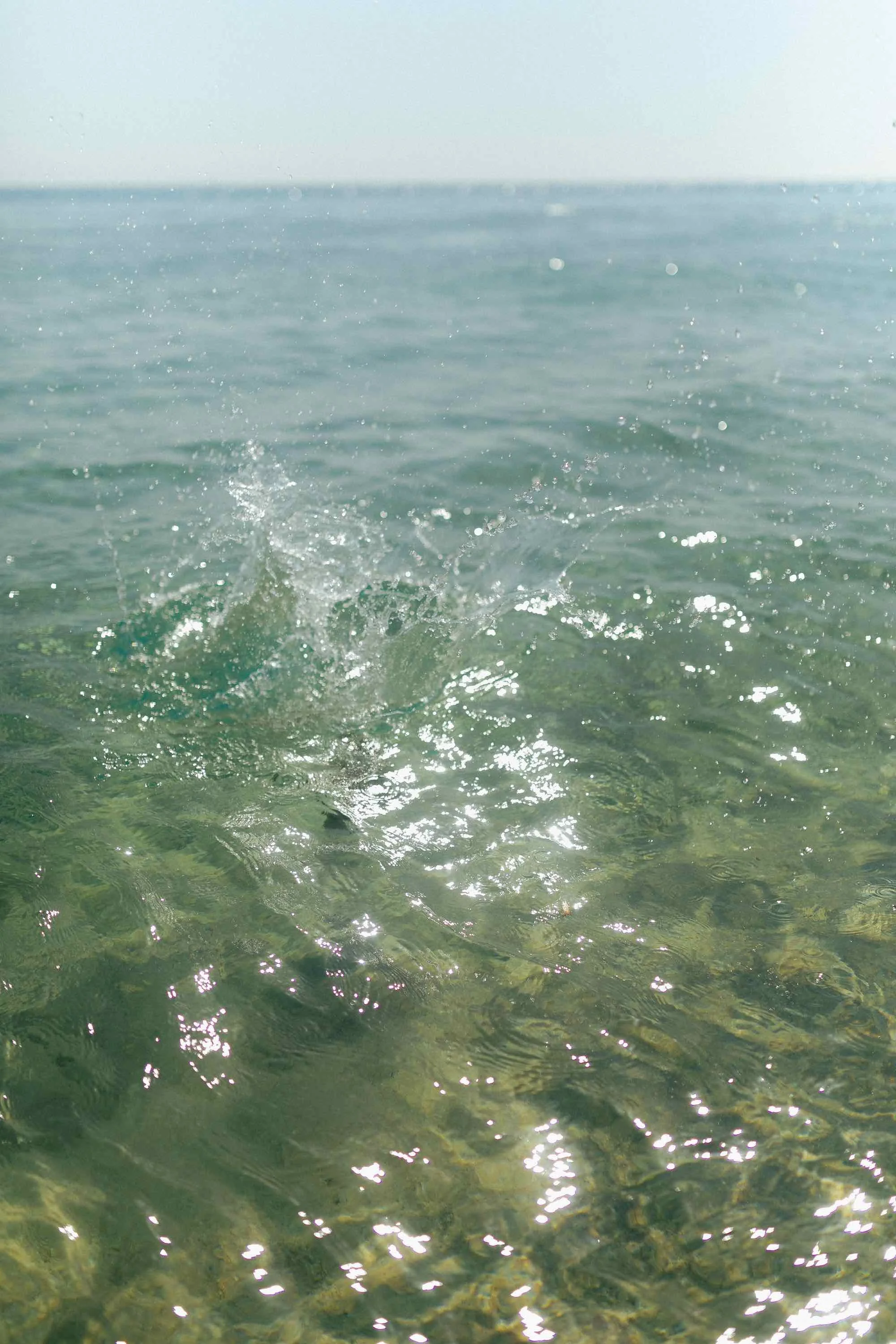Clear ocean water with small splash in the foreground and calm water extending to the horizon under a light blue sky.