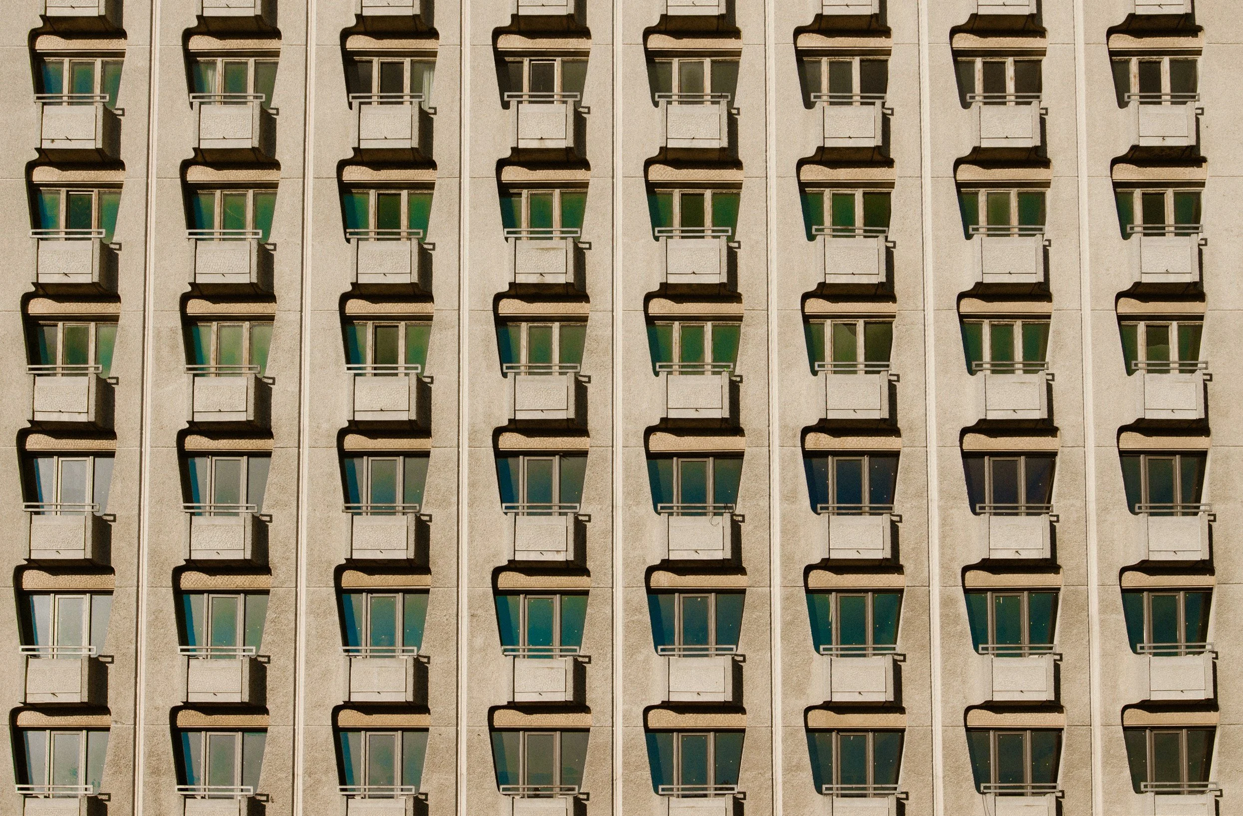 Close-up of a beige-colored building facade with multiple balconies and windows arranged in a grid pattern, casting shadows.