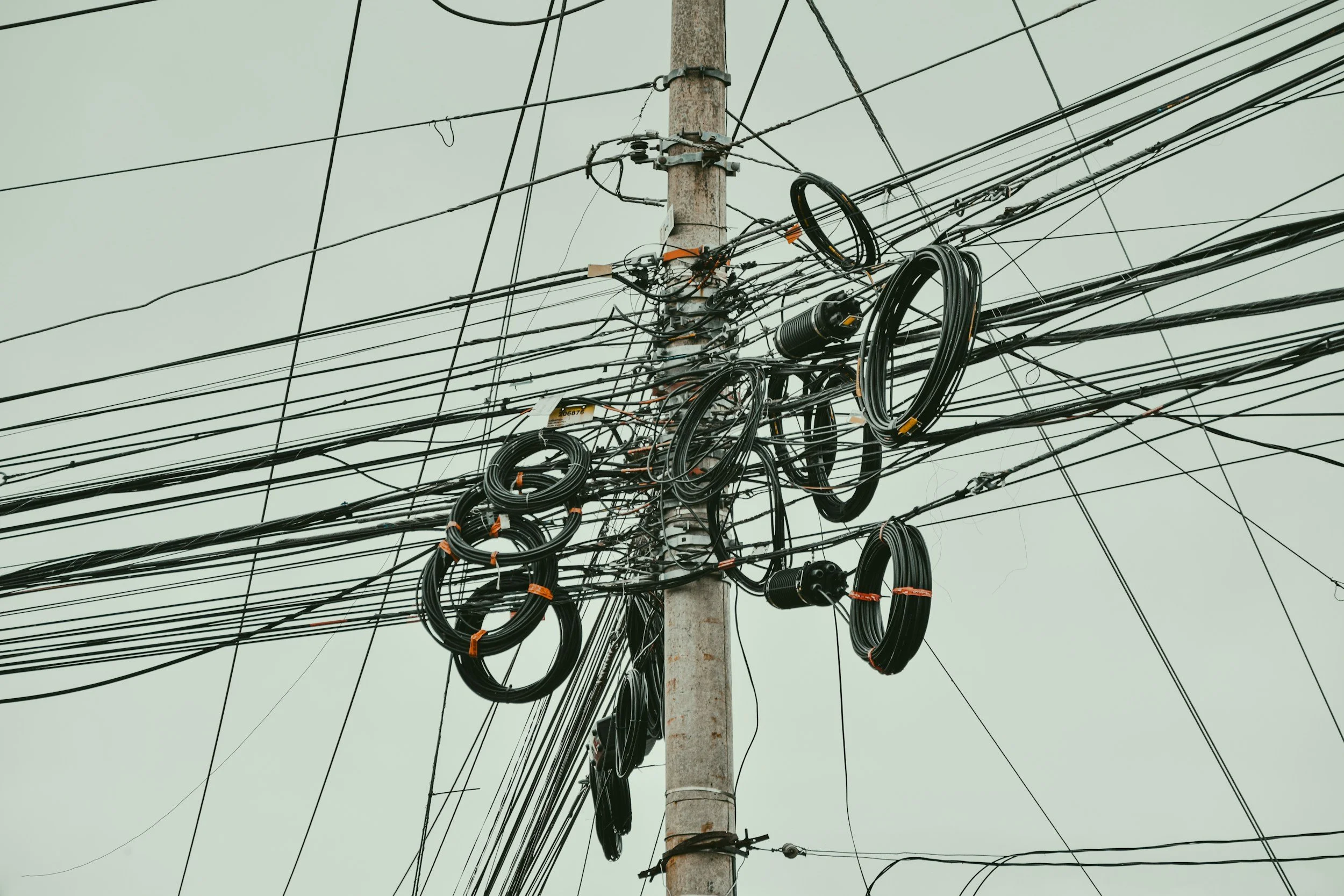 A utility pole with tangled black electrical wires and cables against a pale sky.