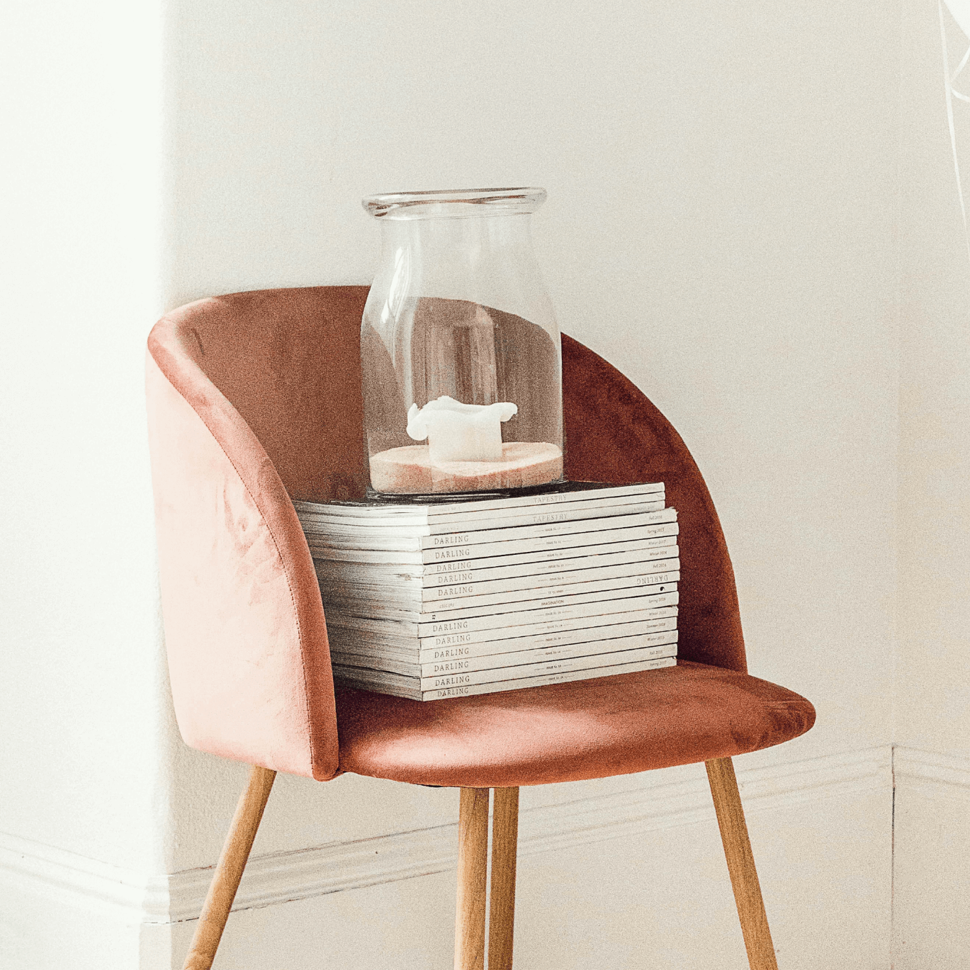A pink velvet chair with wooden legs placed against a plain light-colored wall. On the chair, there is a stack of magazines and a clear glass vase with a white candle inside, positioned on top of the magazines.