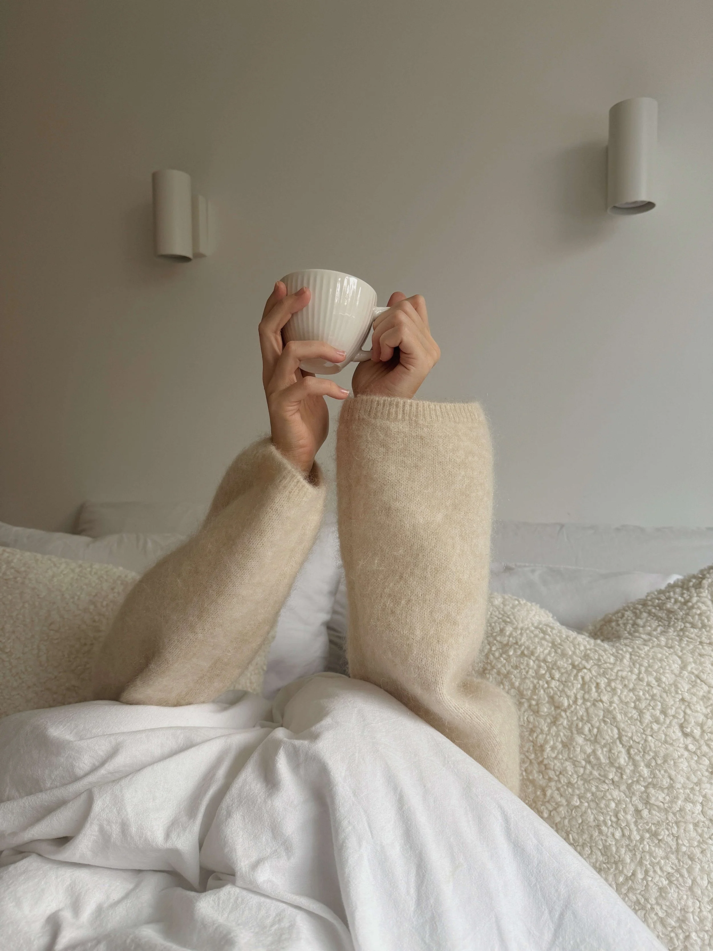 Person in cozy beige sweater holding a white mug while lying in bed with white sheets and textured blankets, against a plain wall with modern white wall lights.