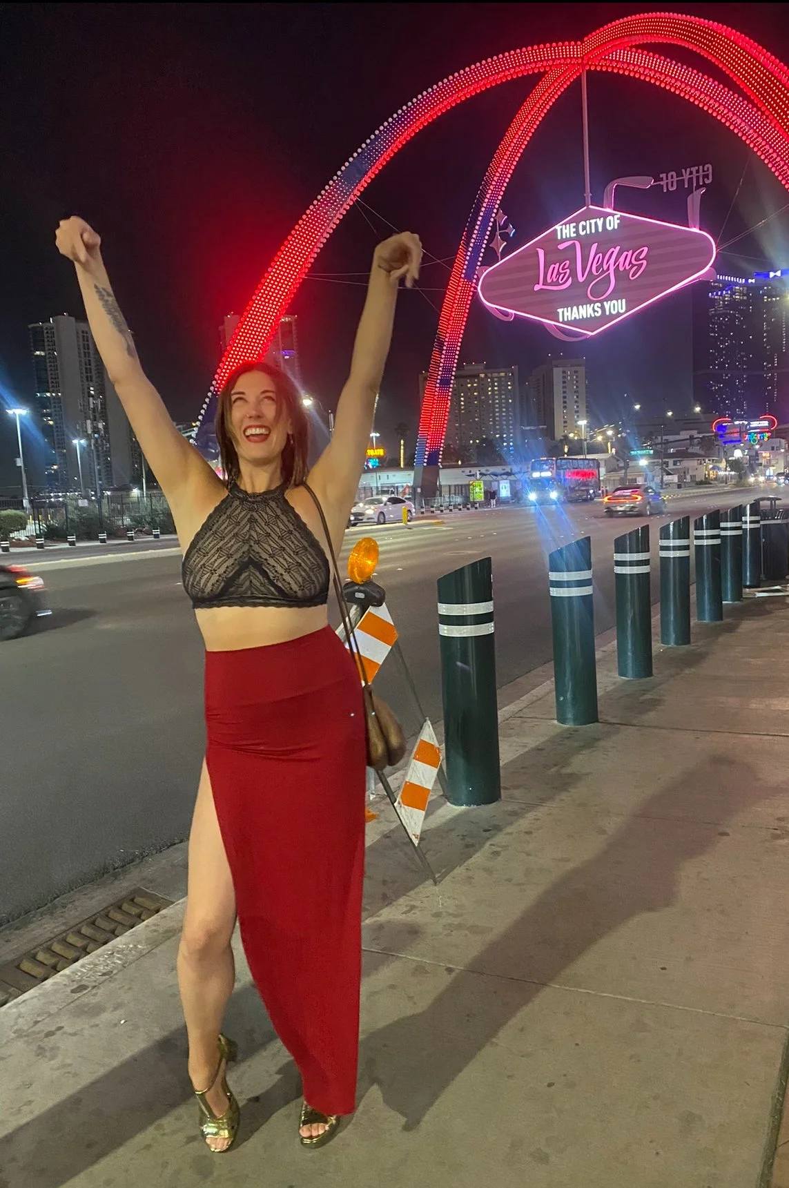 A woman celebrating on the sidewalk in front of the Las Vegas sign at night, with her arms raised and smiling.