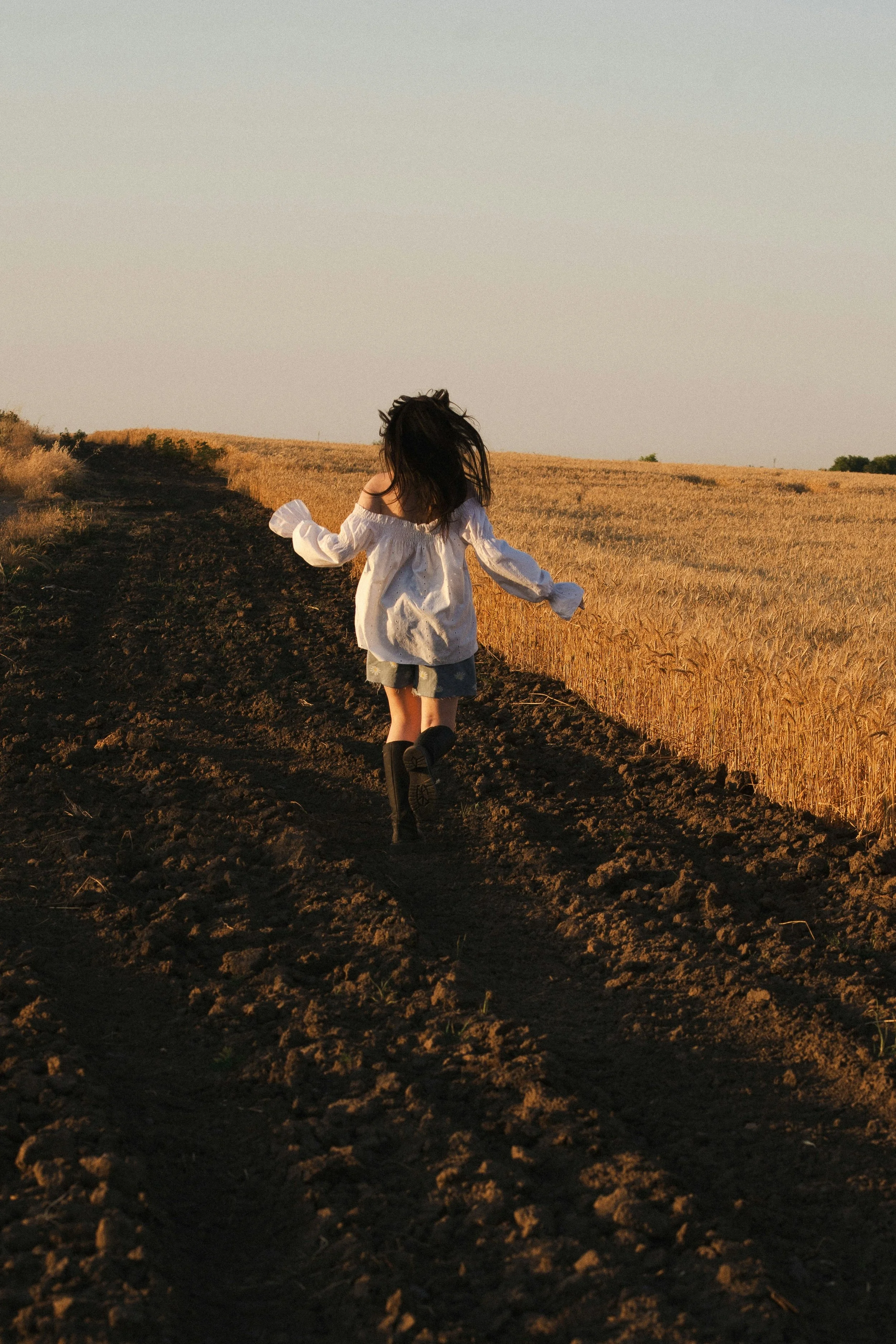 A young girl with dark hair running on a dirt path through a wheat field during sunset.