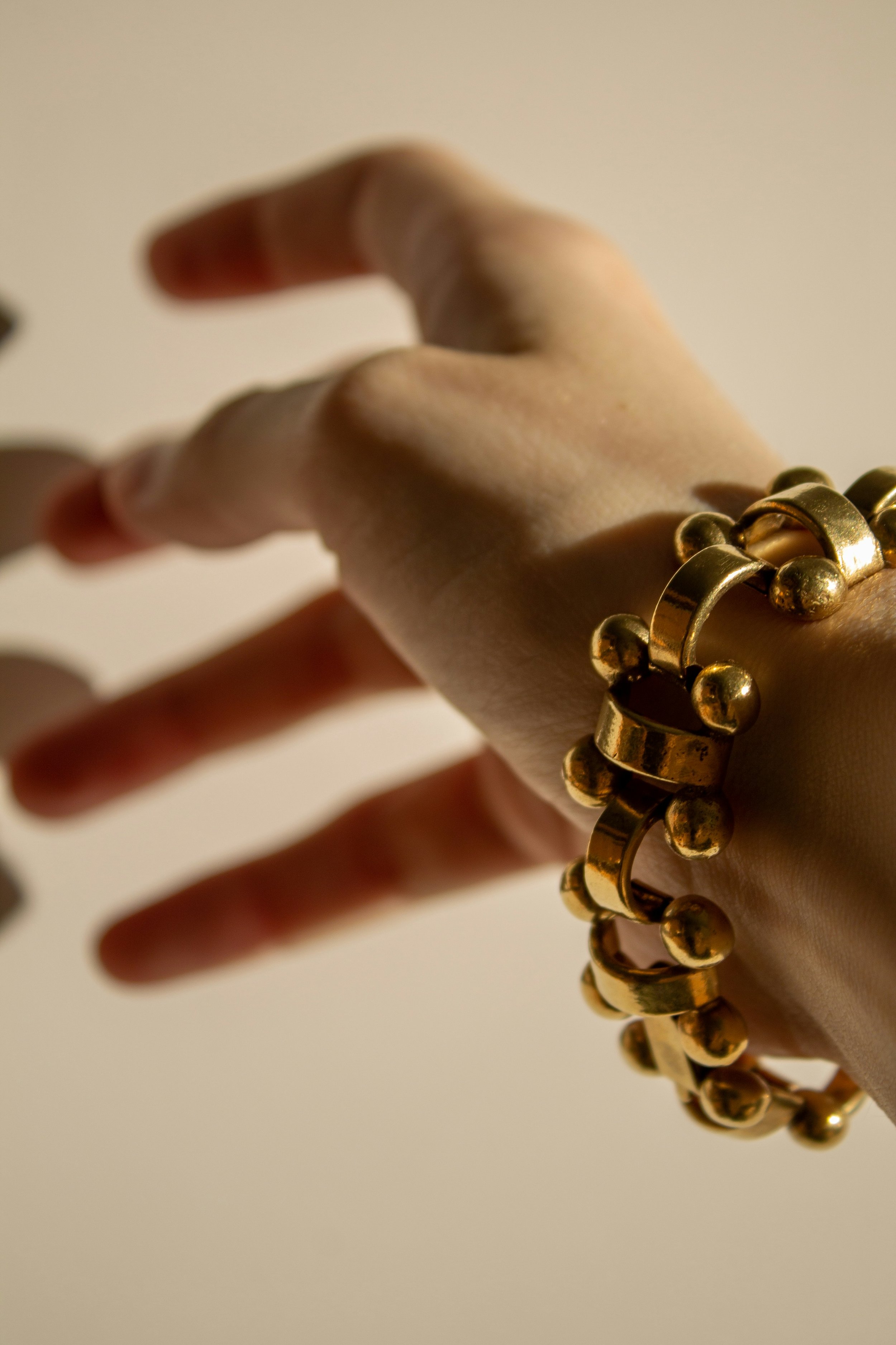Close-up of a hand with gold jewelry, including a chunky bracelet, with fingers slightly spread and shadowed against a plain background.