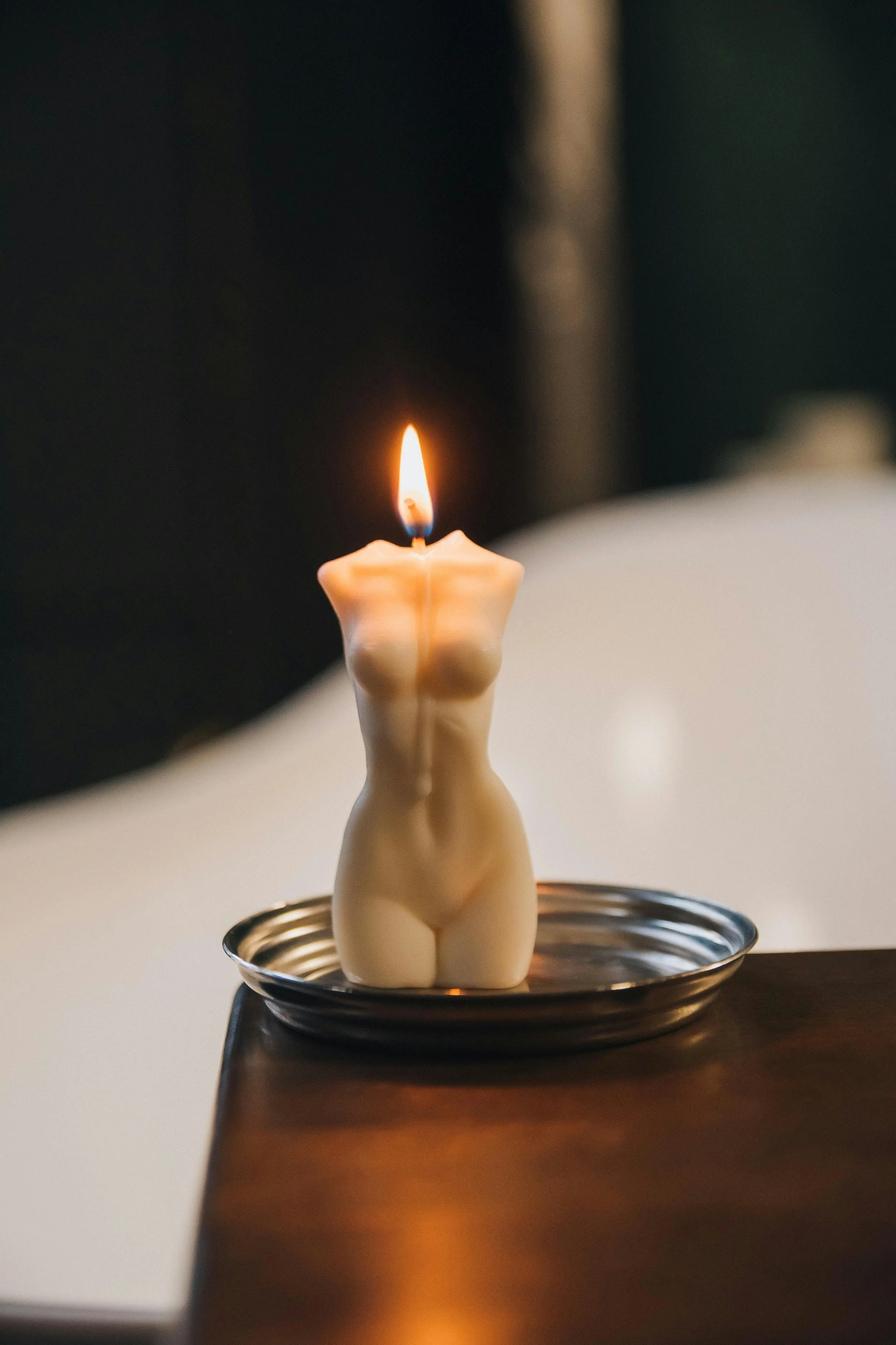 A candle shaped like a female torso is burning, placed on a small metal plate on a wooden surface.