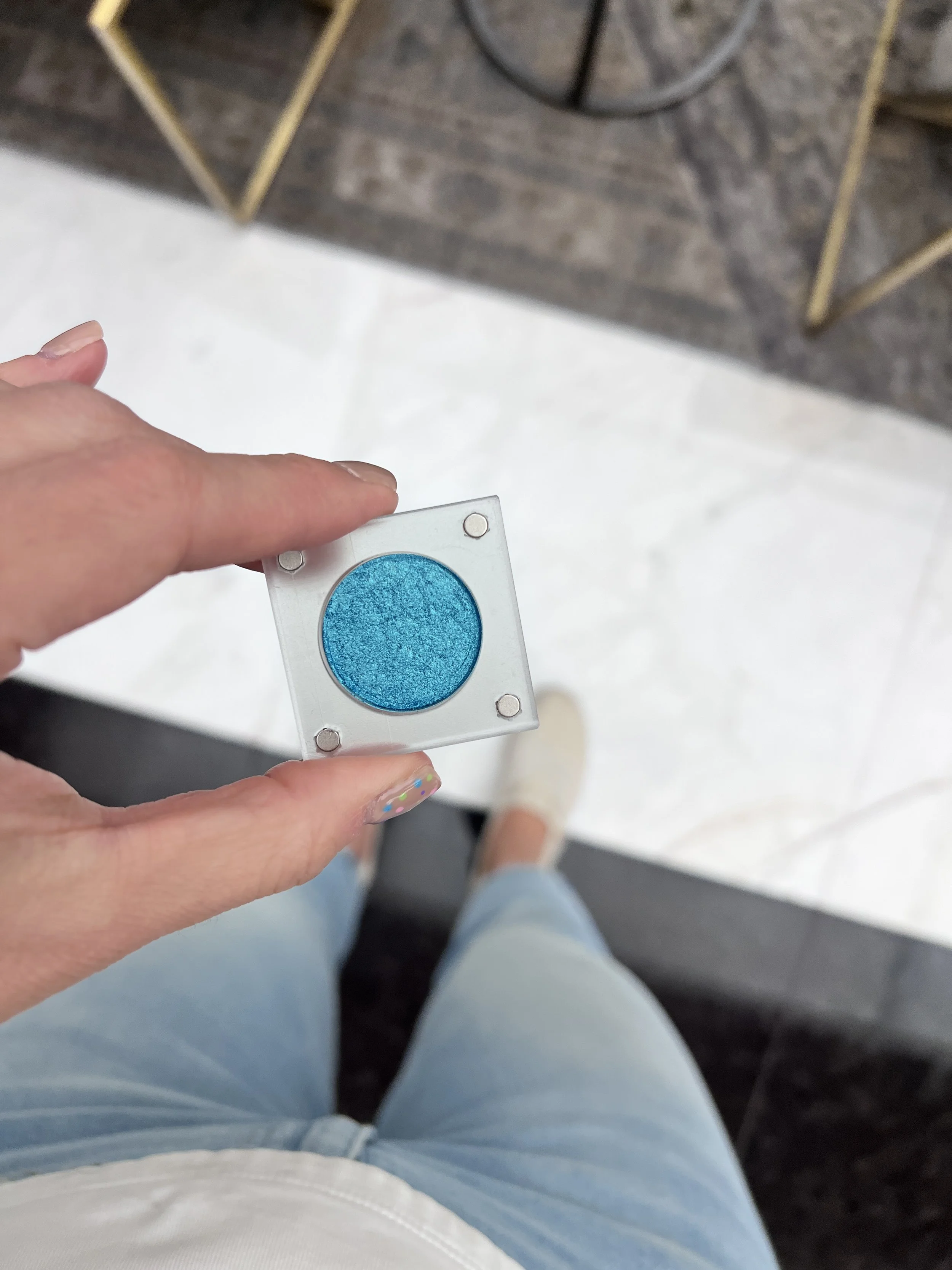 Light-skinned hand holding Breathtaking Glow, a bright blue metallic eyeshadow in a clear magnetic case.