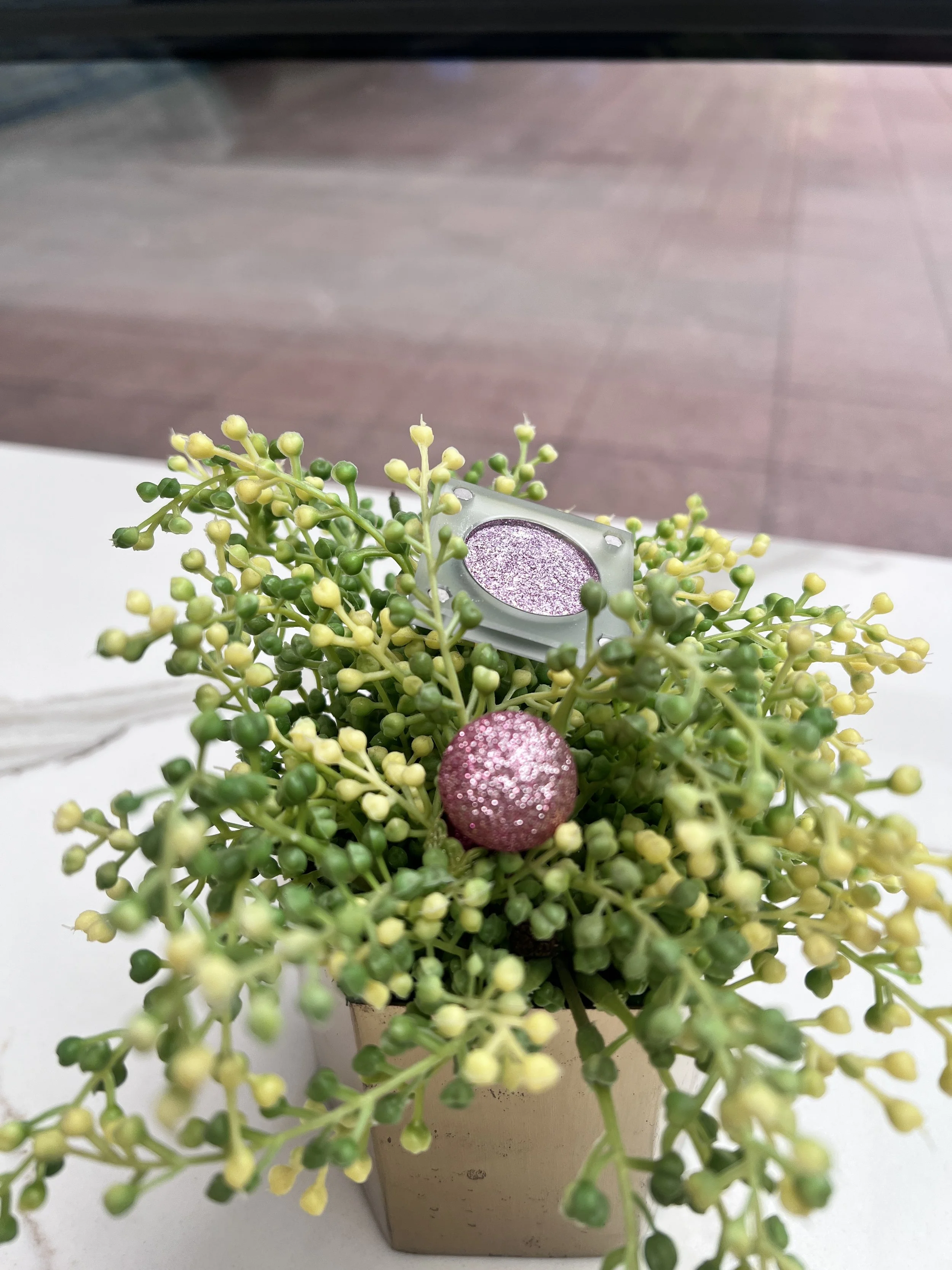Unpredictable Brush, a silvery-lavender glitter eyeshadow, perched in a beaded green plant beside a pink sparkle bauble.