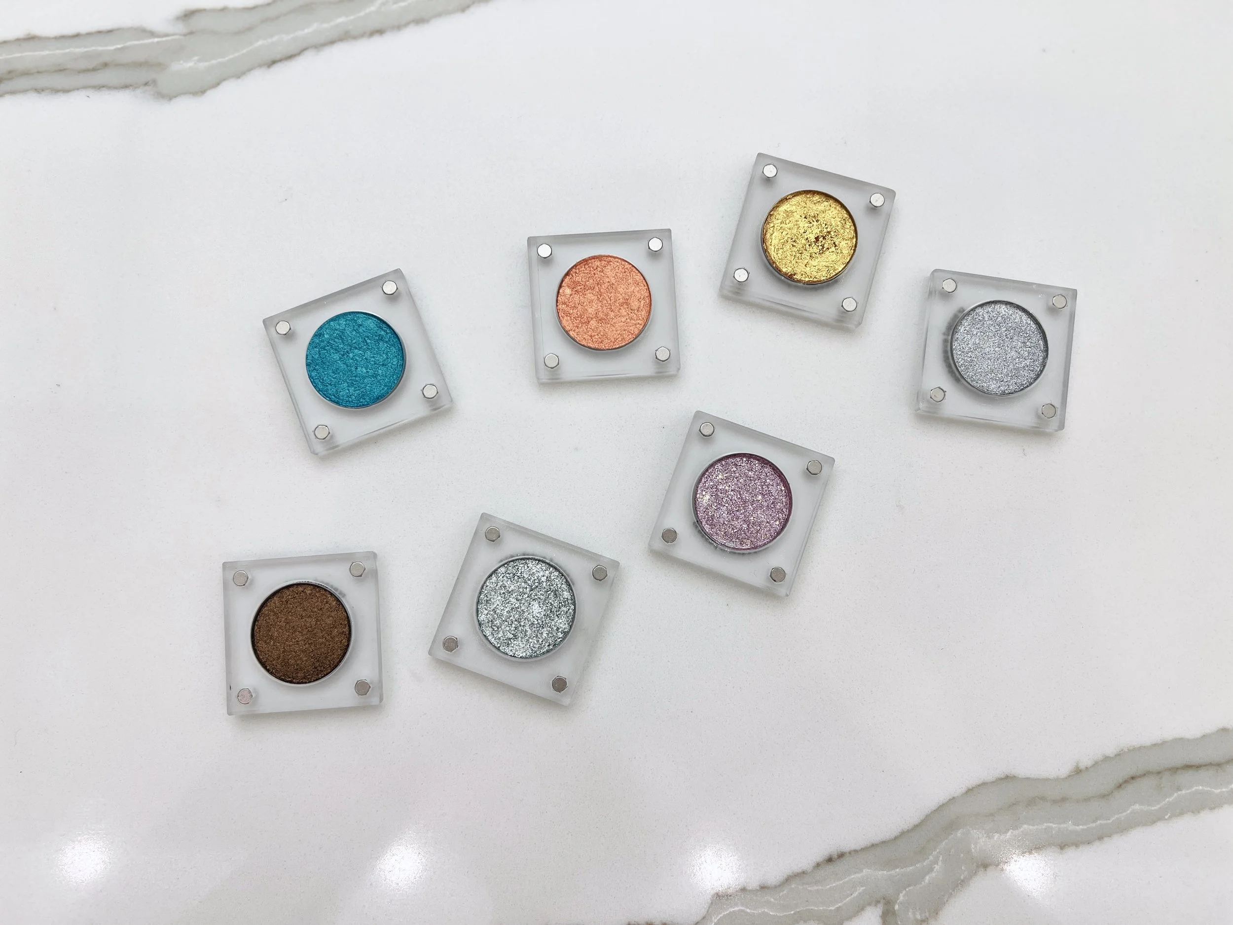 All seven Imperfectionist handmade eyeshadow singles in clear magnetic cases arranged on white marble, including teal, copper, gold, silver, pink glitter, bronze, and icy mint shades.