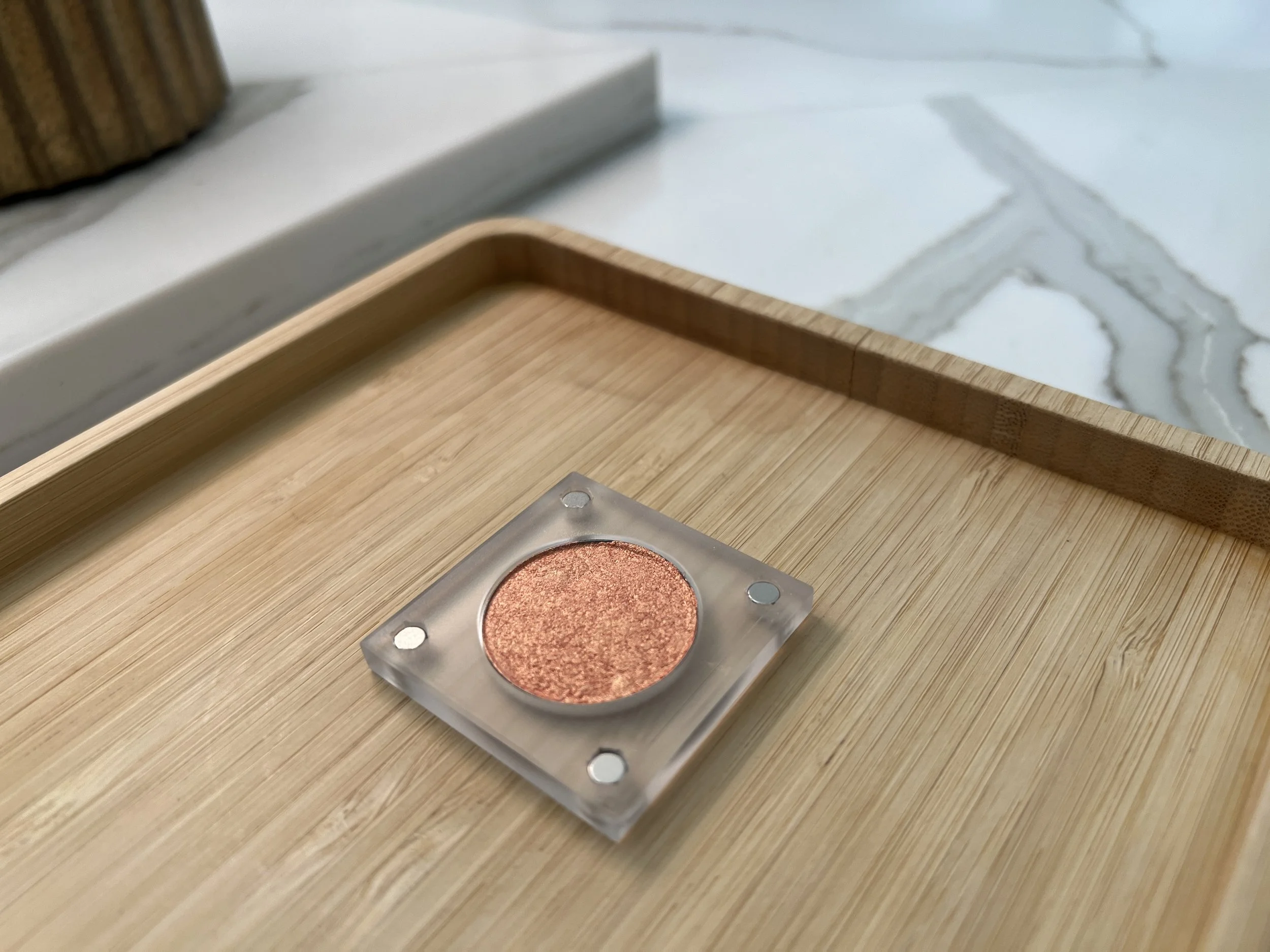 Hands to Crown, a warm rosy-copper eyeshadow with a golden glow, in its clear magnetic case on a bamboo tray.