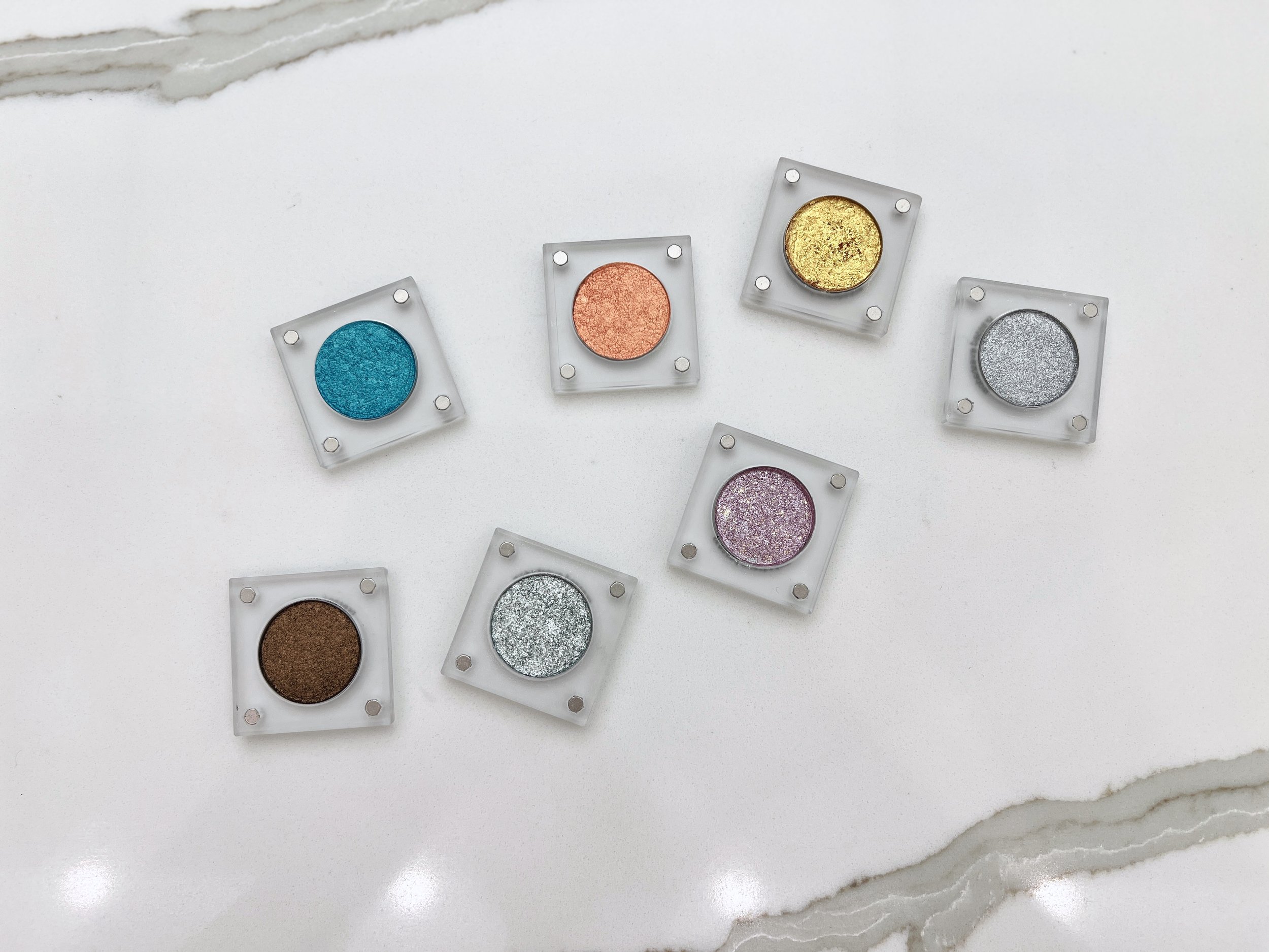 All seven Imperfectionist handmade eyeshadow singles in clear magnetic cases arranged on white marble, including teal, copper, gold, silver, pink glitter, bronze, and icy mint shades.