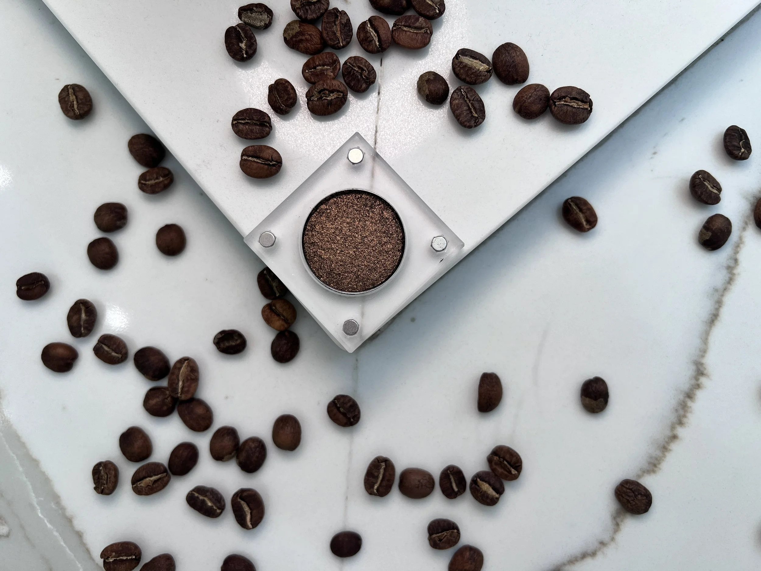 Restless Espresso, a bronze-brown metallic eyeshadow, surrounded by scattered coffee beans on white marble.