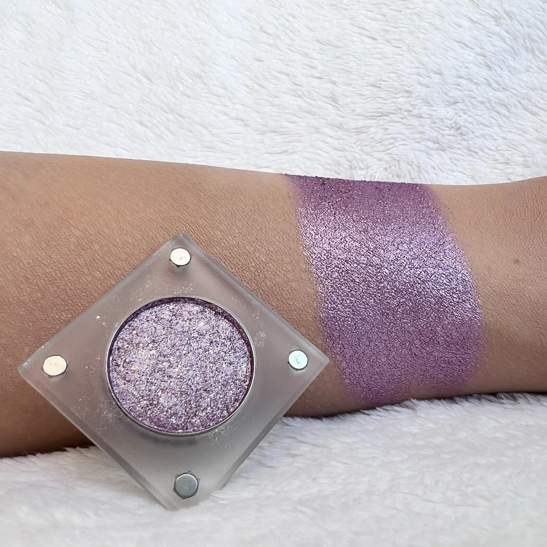 Unpredictable Brush by The Imperfectionist: lilac-purple glitter eyeshadow with sparkle swatched on medium skin next to an open pan.