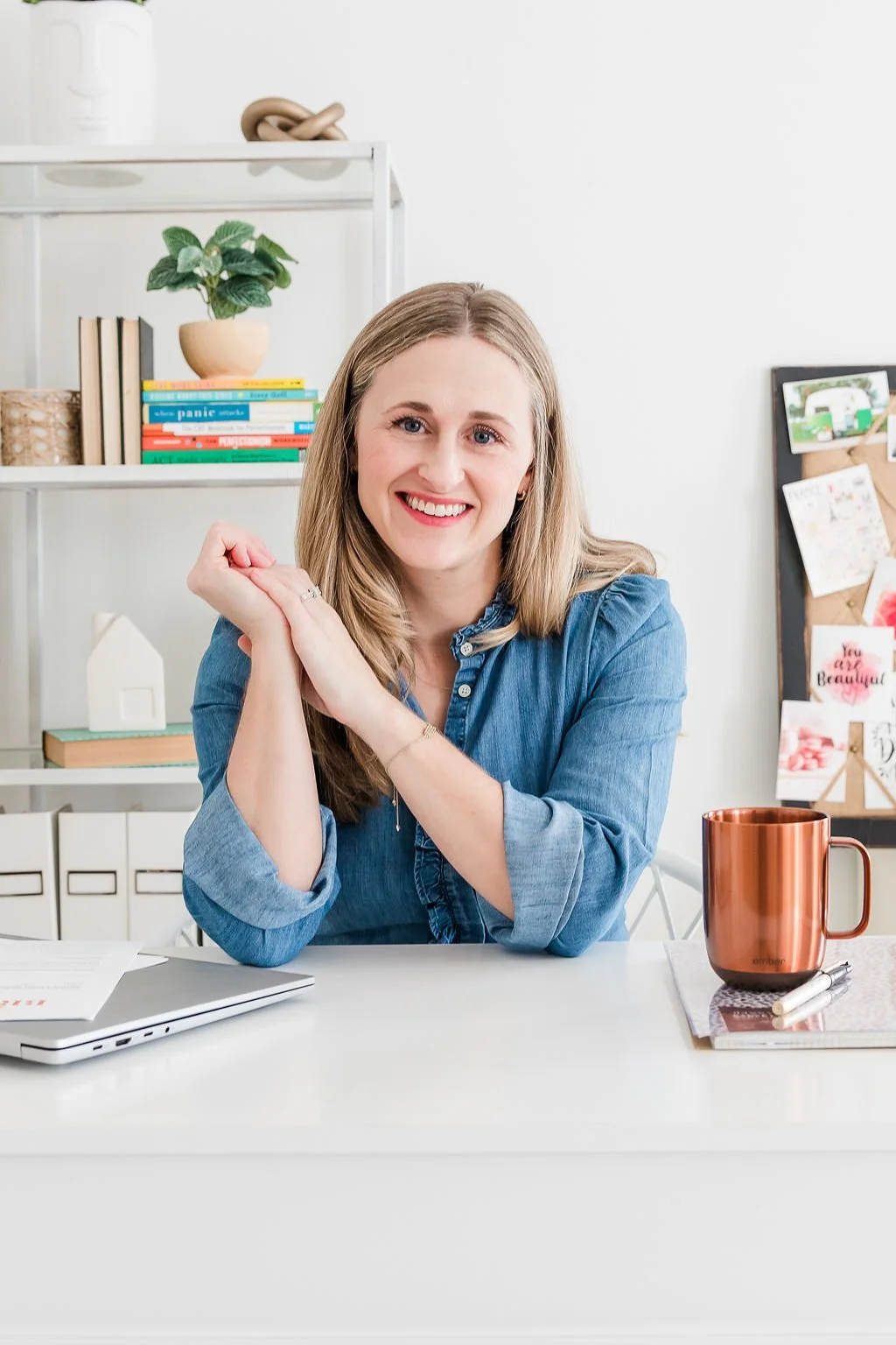 A woman smiling and sitting at a desk in a bright, organized home office with a laptop, a copper mug, and a notebook.