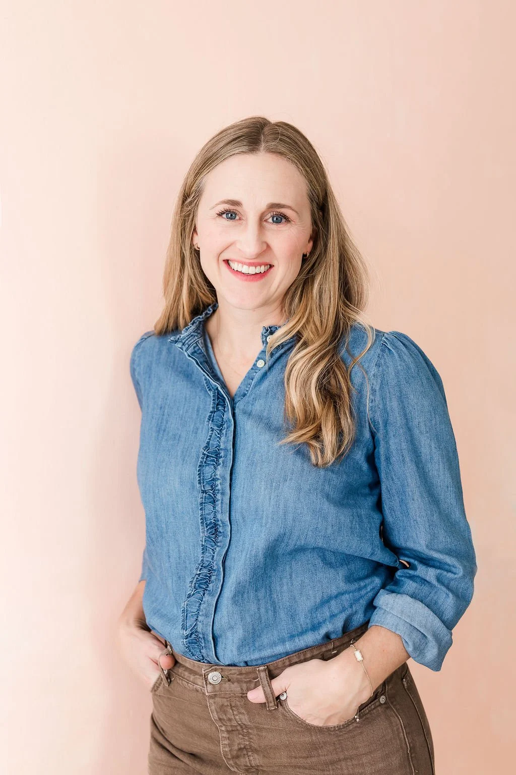 A smiling woman with long blonde hair wearing a denim shirt and brown pants, standing against a light pink background.