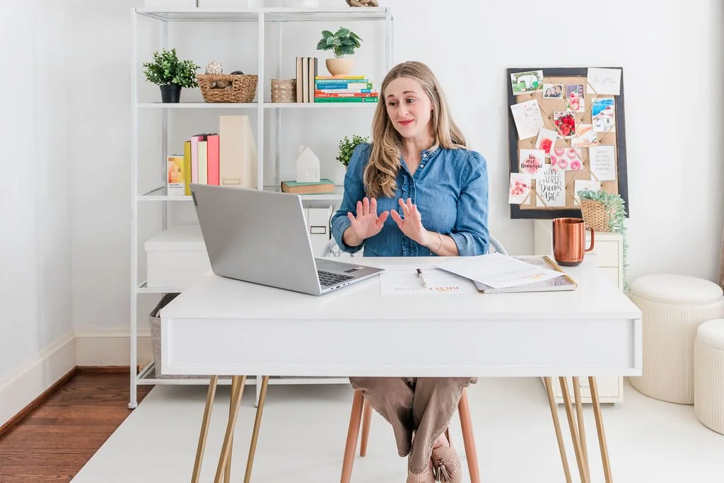 A woman sitting at a white desk with a laptop, papers, and a copper mug, in a bright, organized home office with a white bookshelf and pin board in the background.