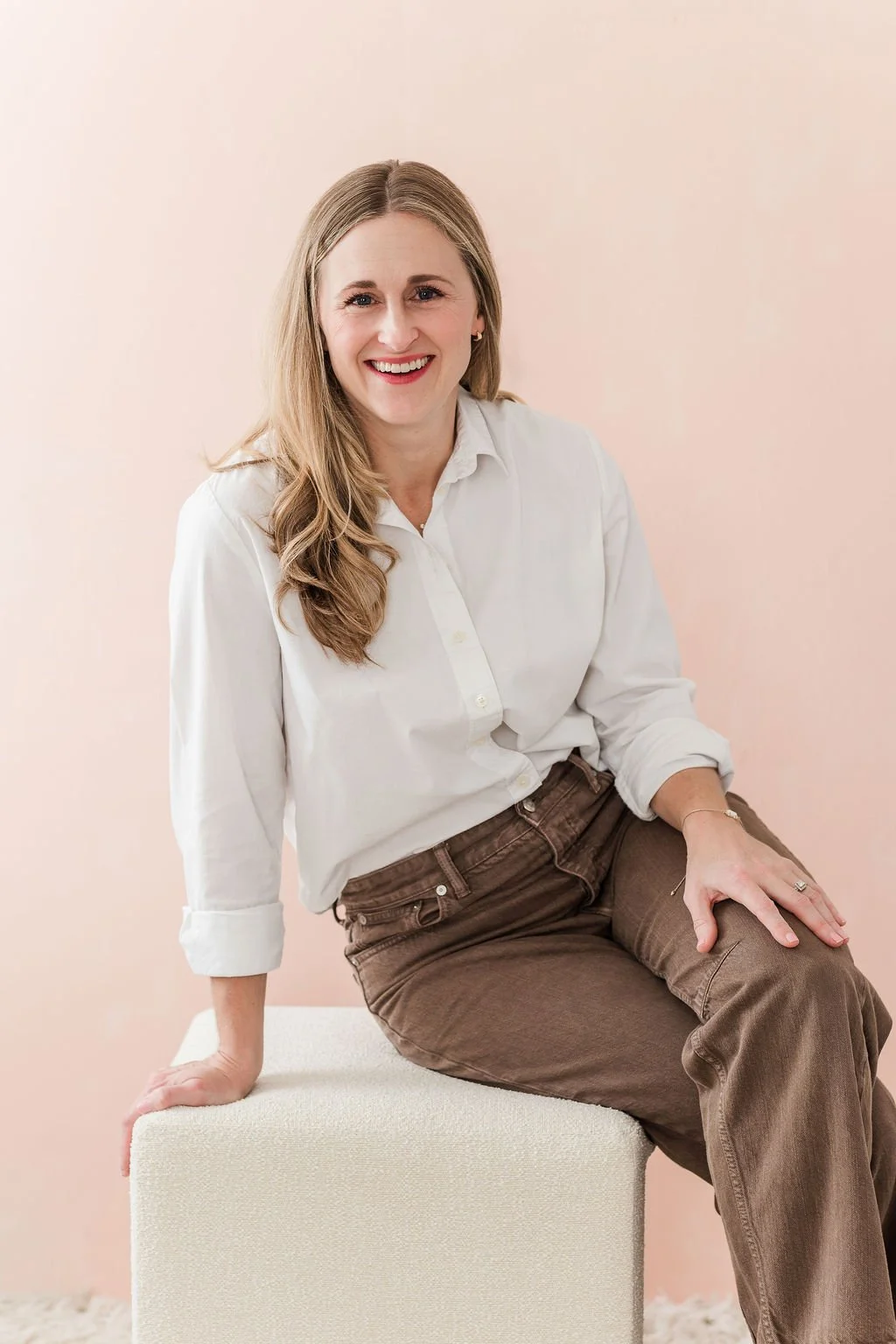A smiling woman with long, wavy blonde hair, wearing a white button-up shirt with sleeves rolled up, and brown pants, sitting on a white textured stool against a plain light pink background.