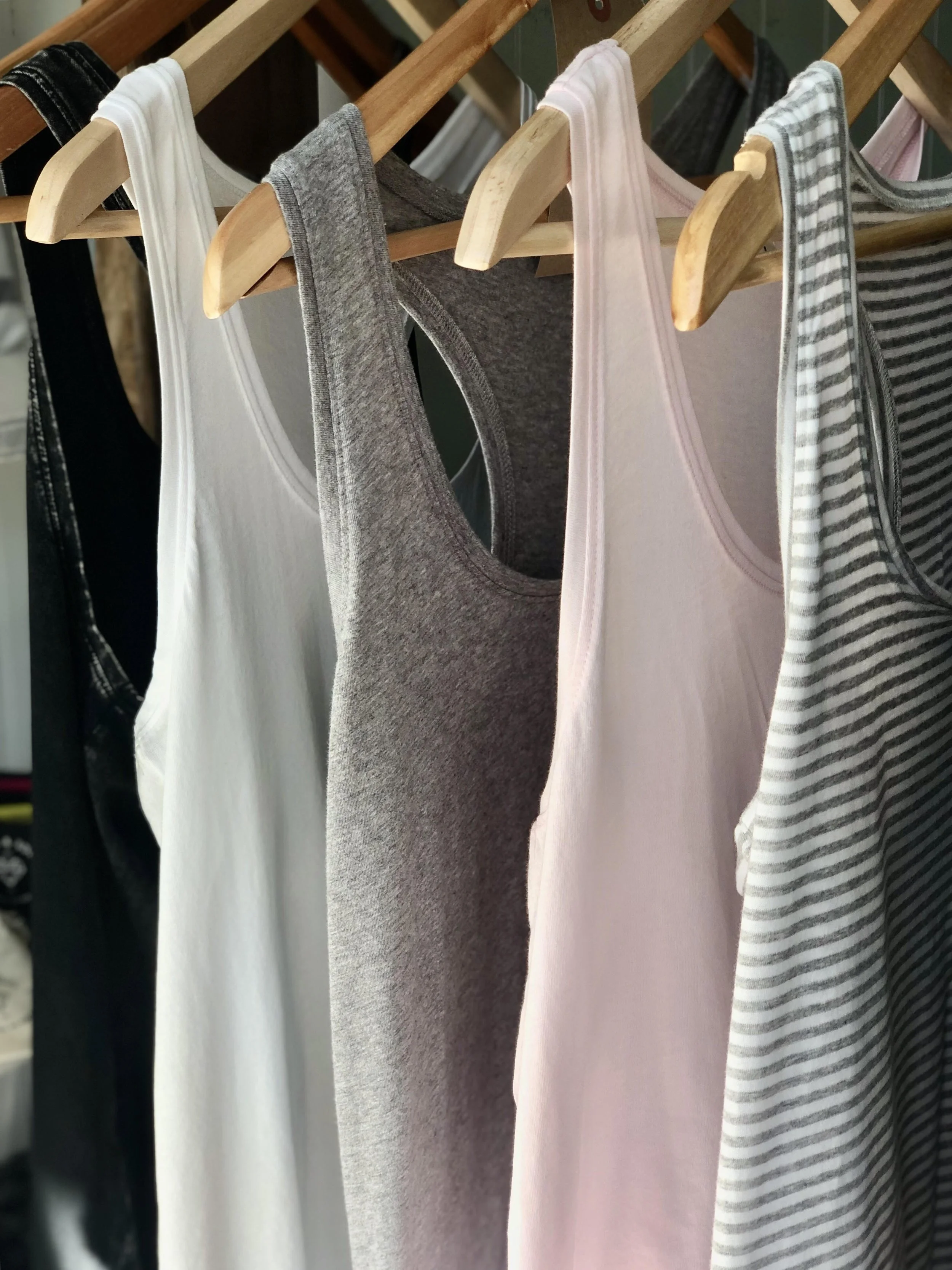 Multiple tank tops hanging on wooden hangers, various colors and patterns including black, white, gray, pink, and striped, on a wooden clothing rack.