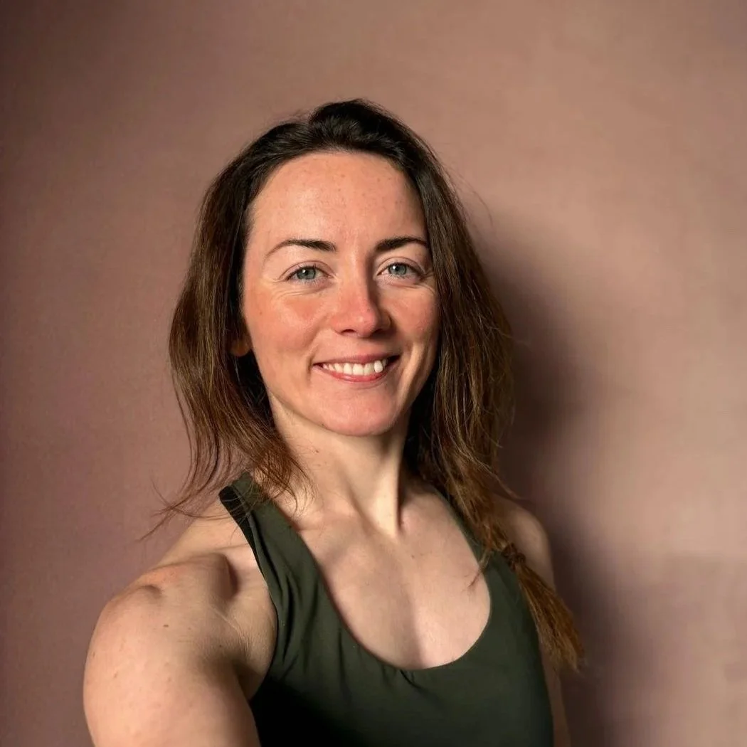 A woman with shoulder-length brown hair and blue eyes smiling, wearing a dark green sleeveless top, standing against a plain brown background.