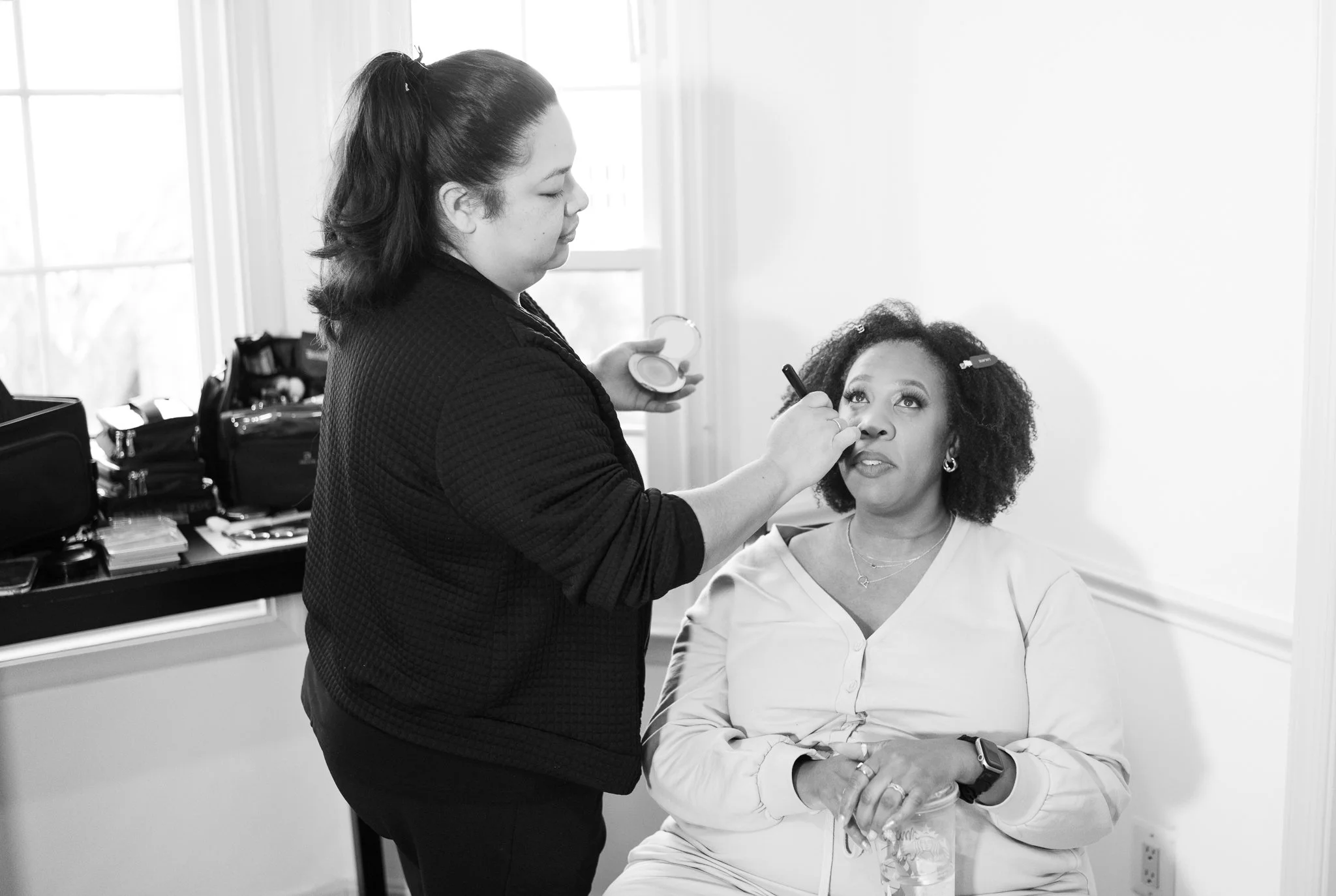 Makeup artist applying makeup on woman seated in a room with natural light.