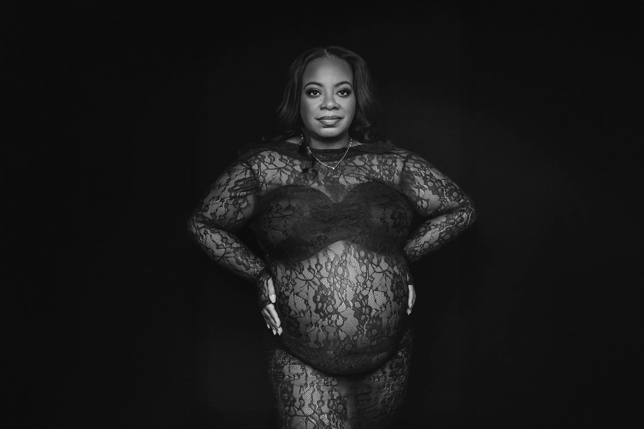 A pregnant woman in a sheer black lace dress with long sleeves poses against a black background, looking confidently at the camera.