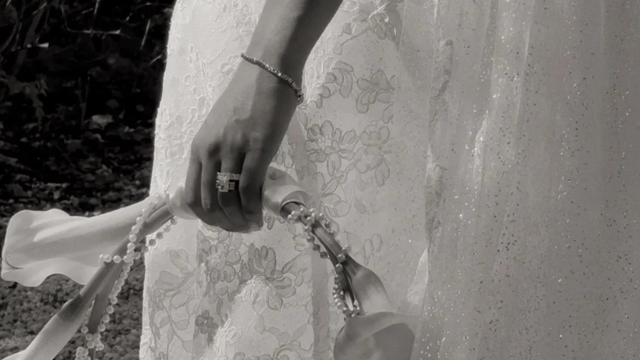 Close-up of a hand pouring a liquid onto a surface, with jewelry including a ring and bracelet, holding a spoon with a chain, and a lace fabric in the background, all shown in black and white.