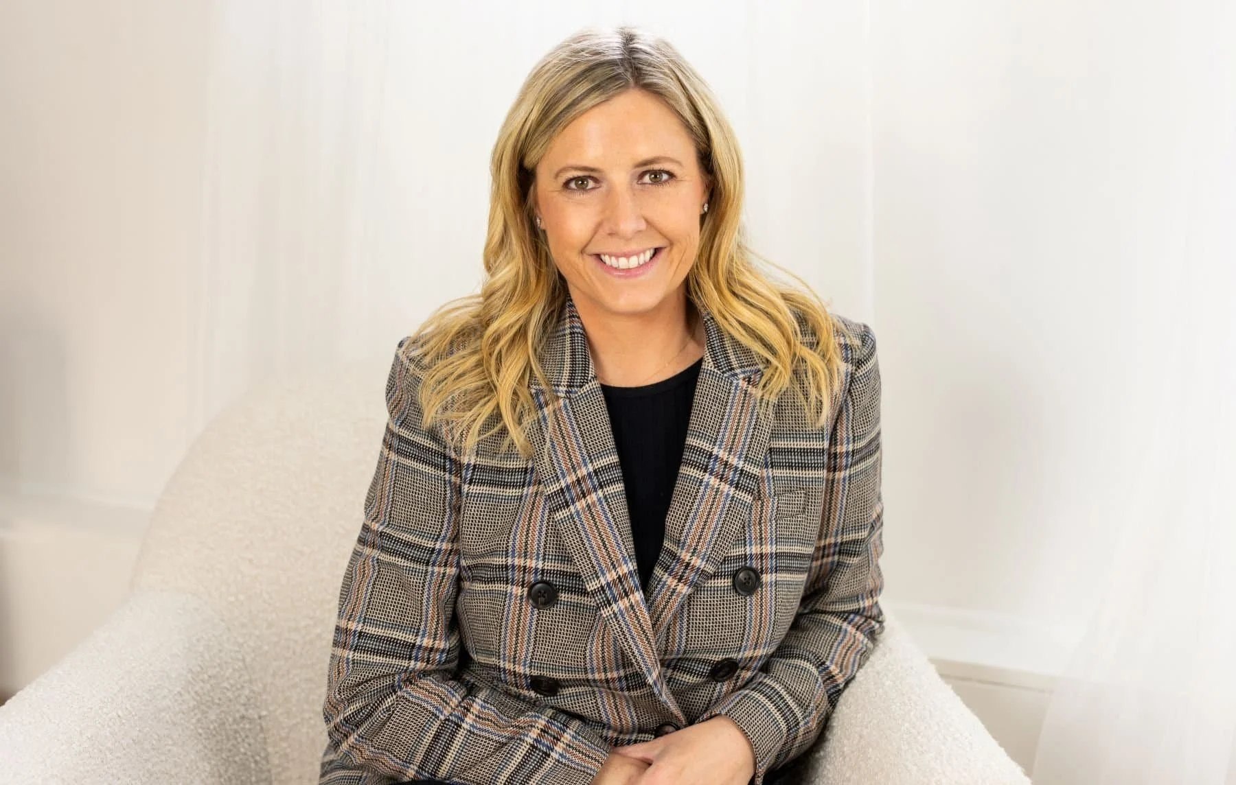 A woman with blonde wavy hair smiling, wearing a plaid blazer and sitting on a light-colored chair in a bright room.