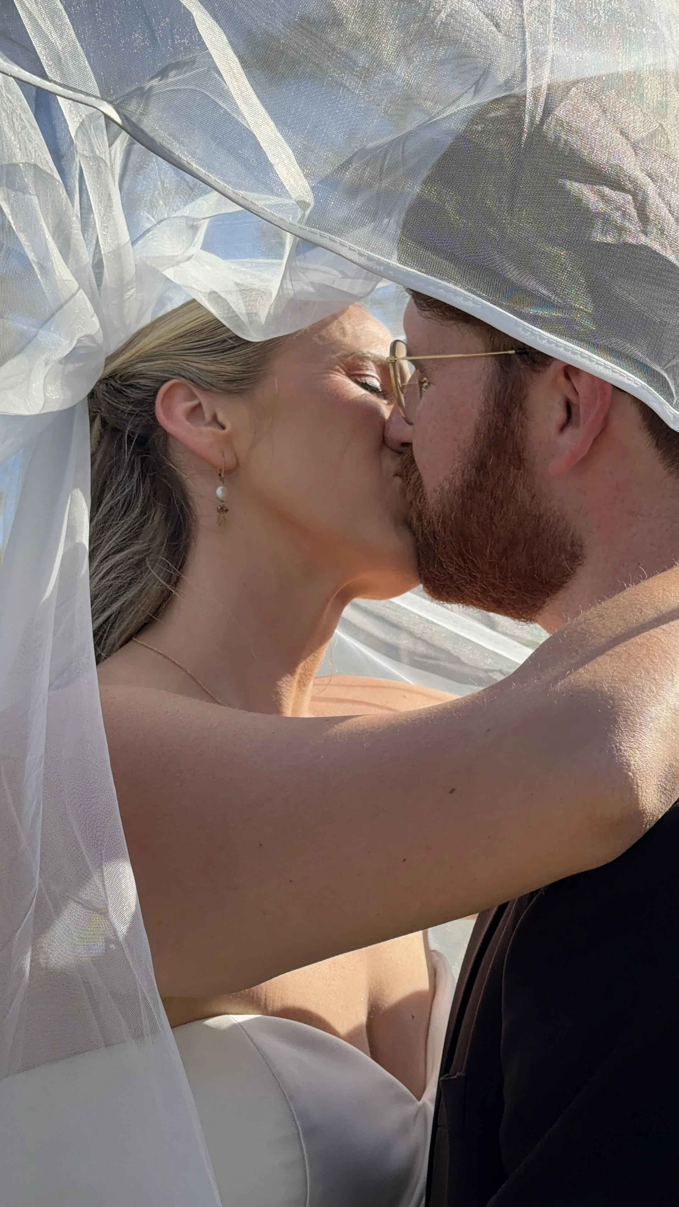 A couple sharing a kiss outdoors, with the woman wearing a strapless white dress and the man wearing glasses and a black suit. The woman has jewelry, including earrings and a necklace, and a sheer veil is draped over them.