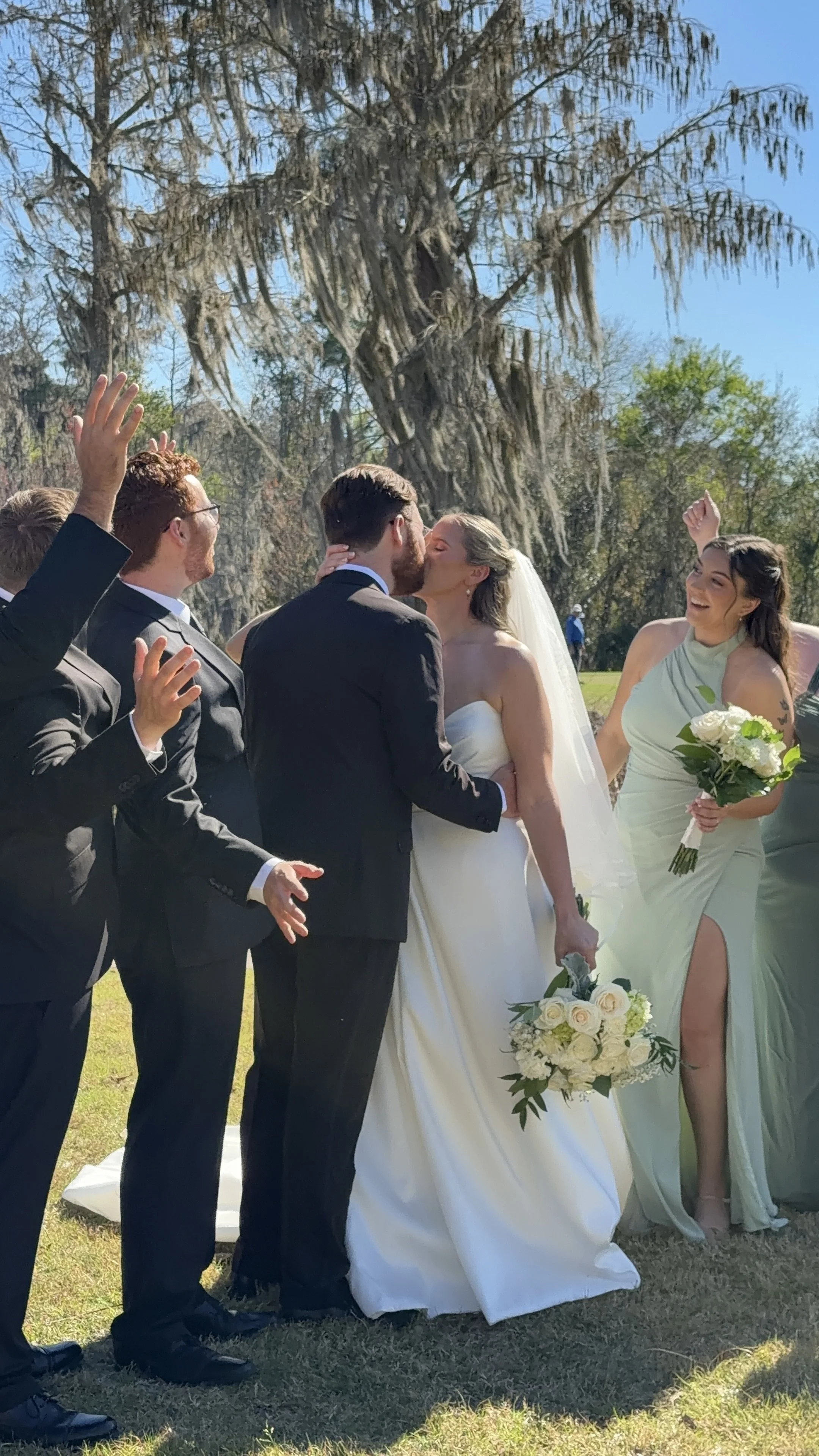 A wedding ceremony outdoors with a bride and groom kissing, surrounded by friends in formal attire, celebrating in front of a large tree with Spanish moss on its branches.