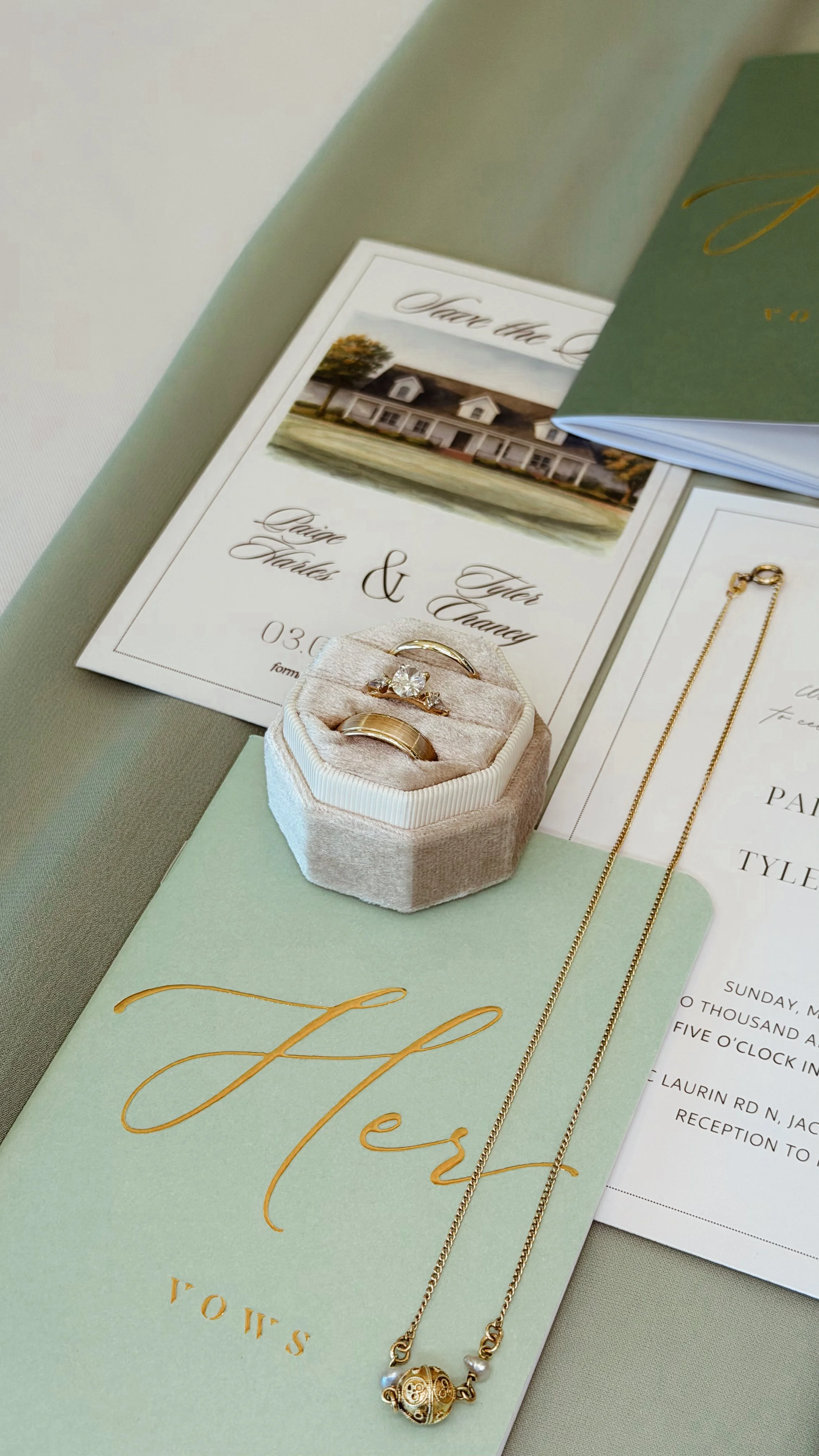 Wedding invitation cards, gold jewelry including a necklace and rings, and a white ring box with a diamond ring on an elegant table.