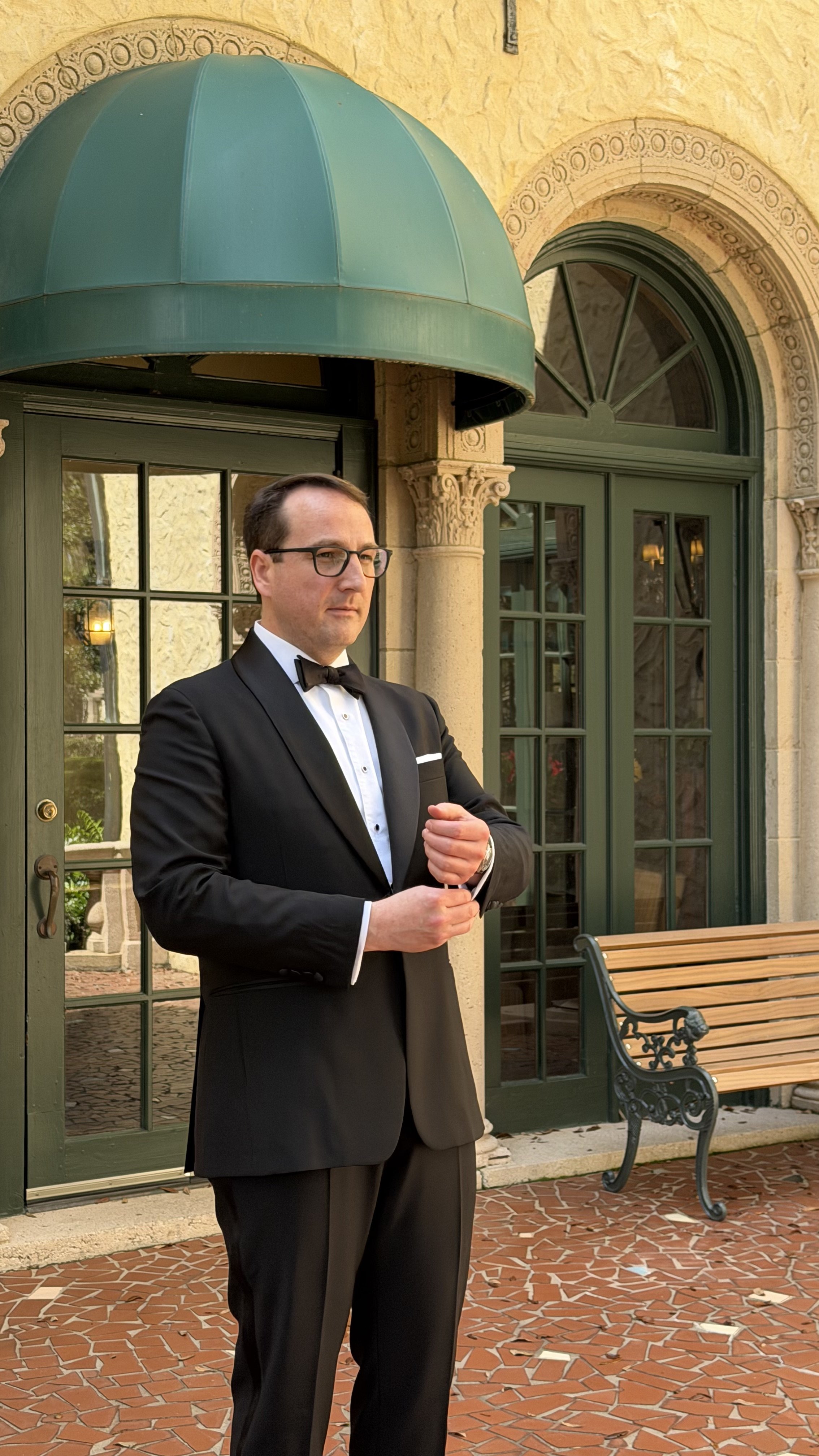 A man in a formal tuxedo with a bow tie stands outdoors in front of a building with green doors and windows, a decorative archway, and a green awning.