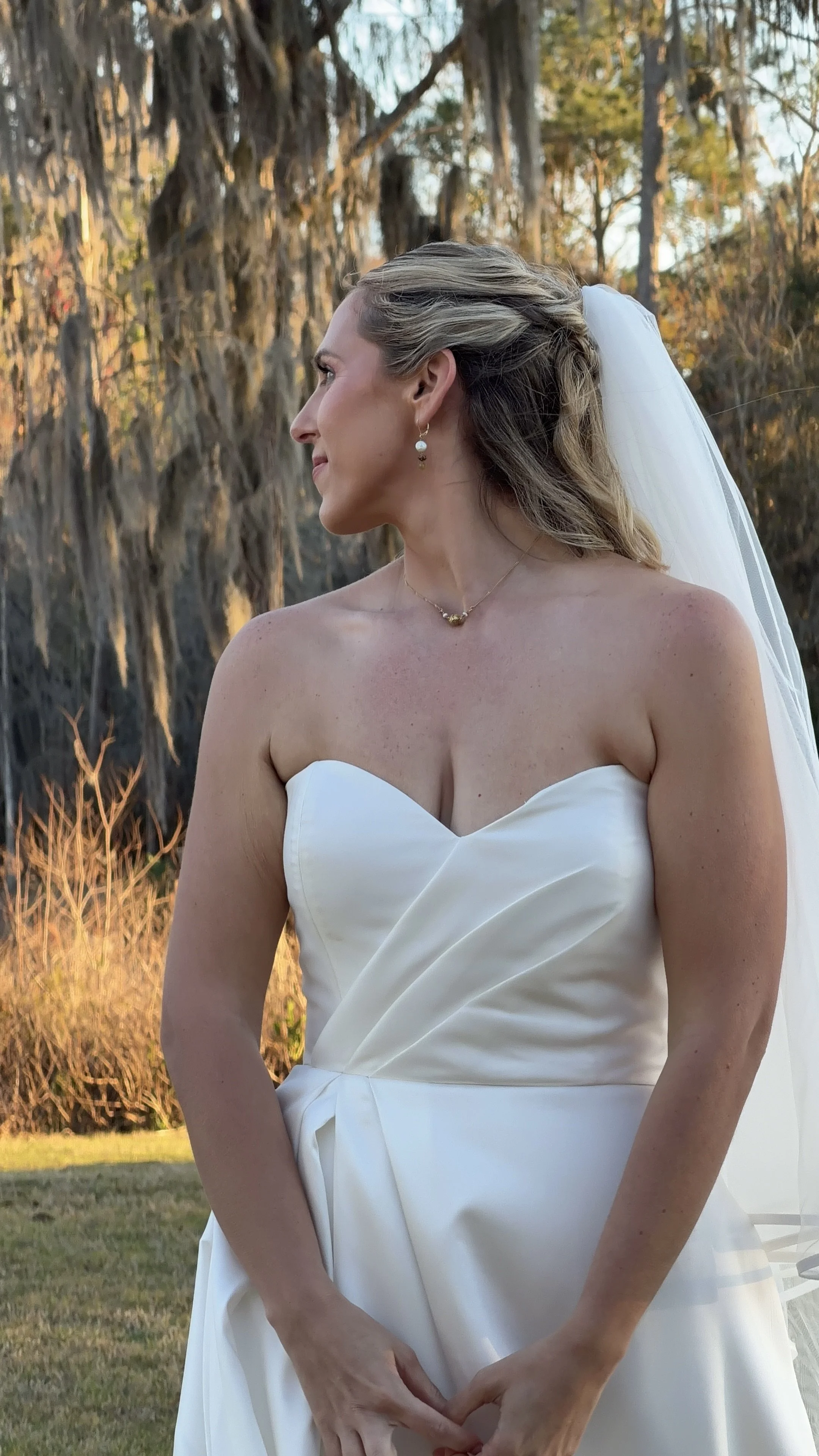 A bride in a strapless white wedding gown with a veil, standing outdoors during sunset, with trees and grass in the background.