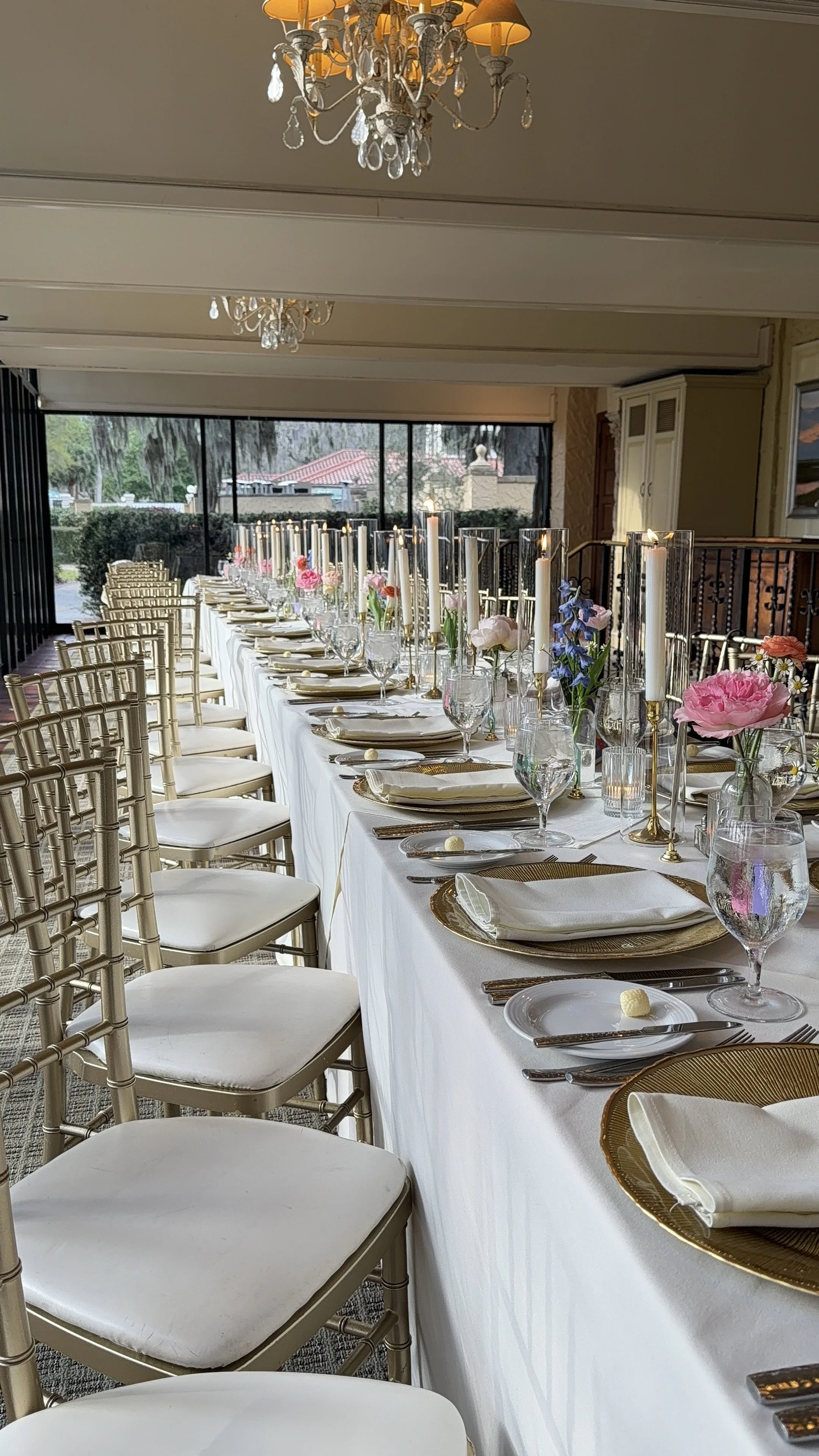 Elegant dining table set for a formal event with white tablecloth, gold-rimmed plates, napkins, glassware, gold chairs, floral centerpieces, and tall candles under a chandelier.