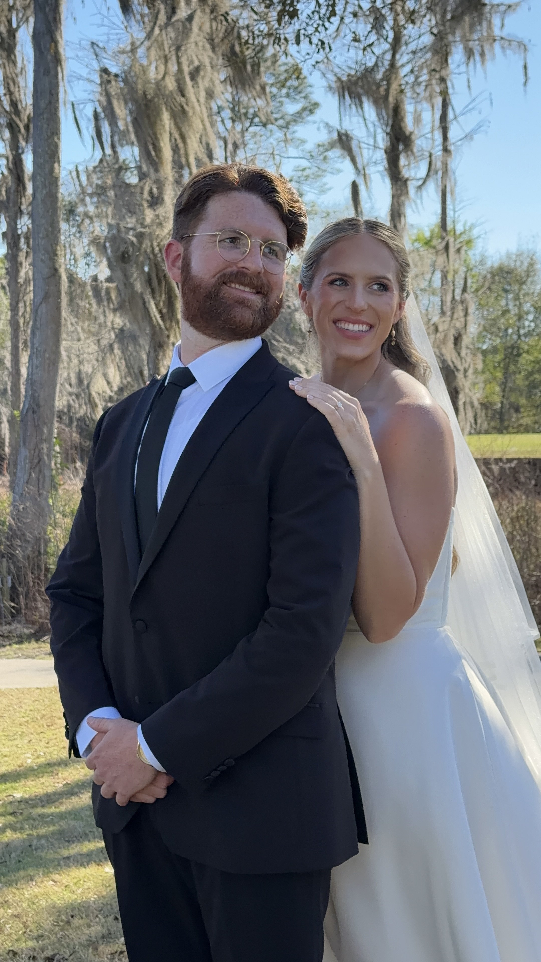 A bride and groom posing outdoors on their wedding day, with trees in the background. The groom is wearing a black suit with a white shirt and black tie, and the bride is wearing a white wedding dress and veil, smiling.